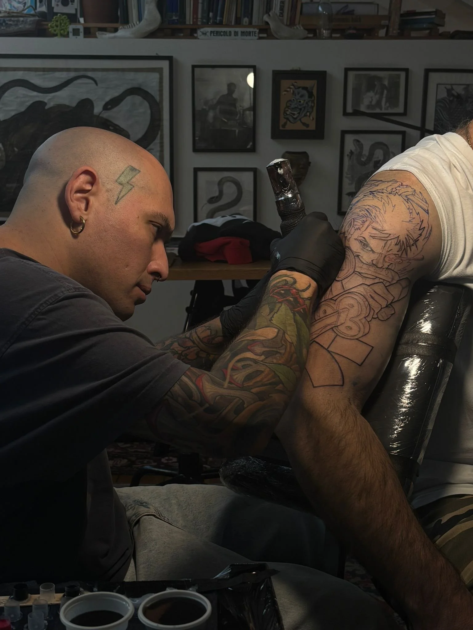 A tattoo artist with colorful tattoos on his arms tattooing a person's arm with a line art design of a character on their upper arm in a tattoo studio. The background features framed artwork on the wall.