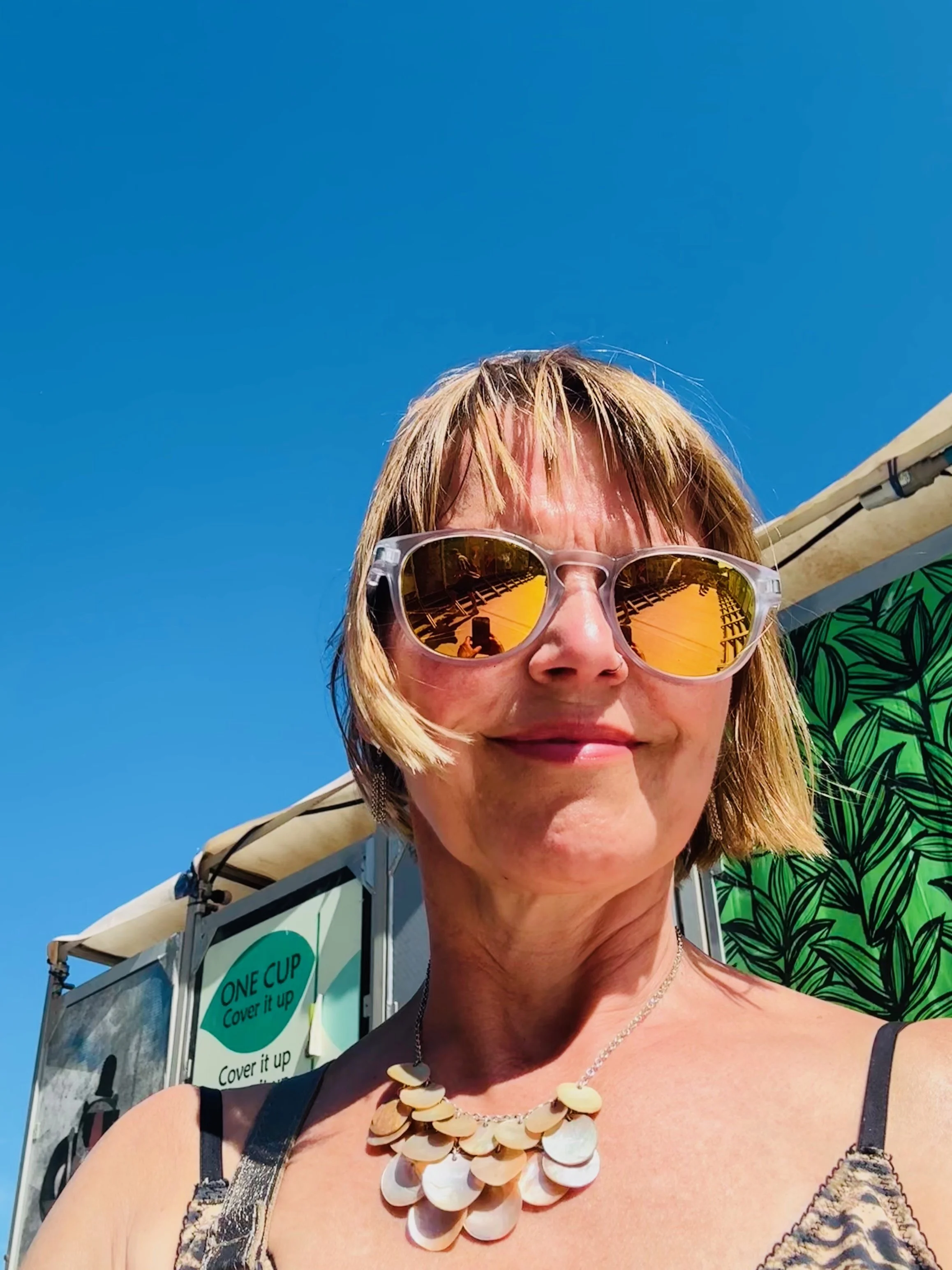 Woman with short blonde hair wearing sunglasses with orange reflections, a shell necklace, and a tank top, standing outdoors under a bright blue sky. A booth with green leafy design and a sign reading 'ONE CUP Cover it up' is visible behind her.