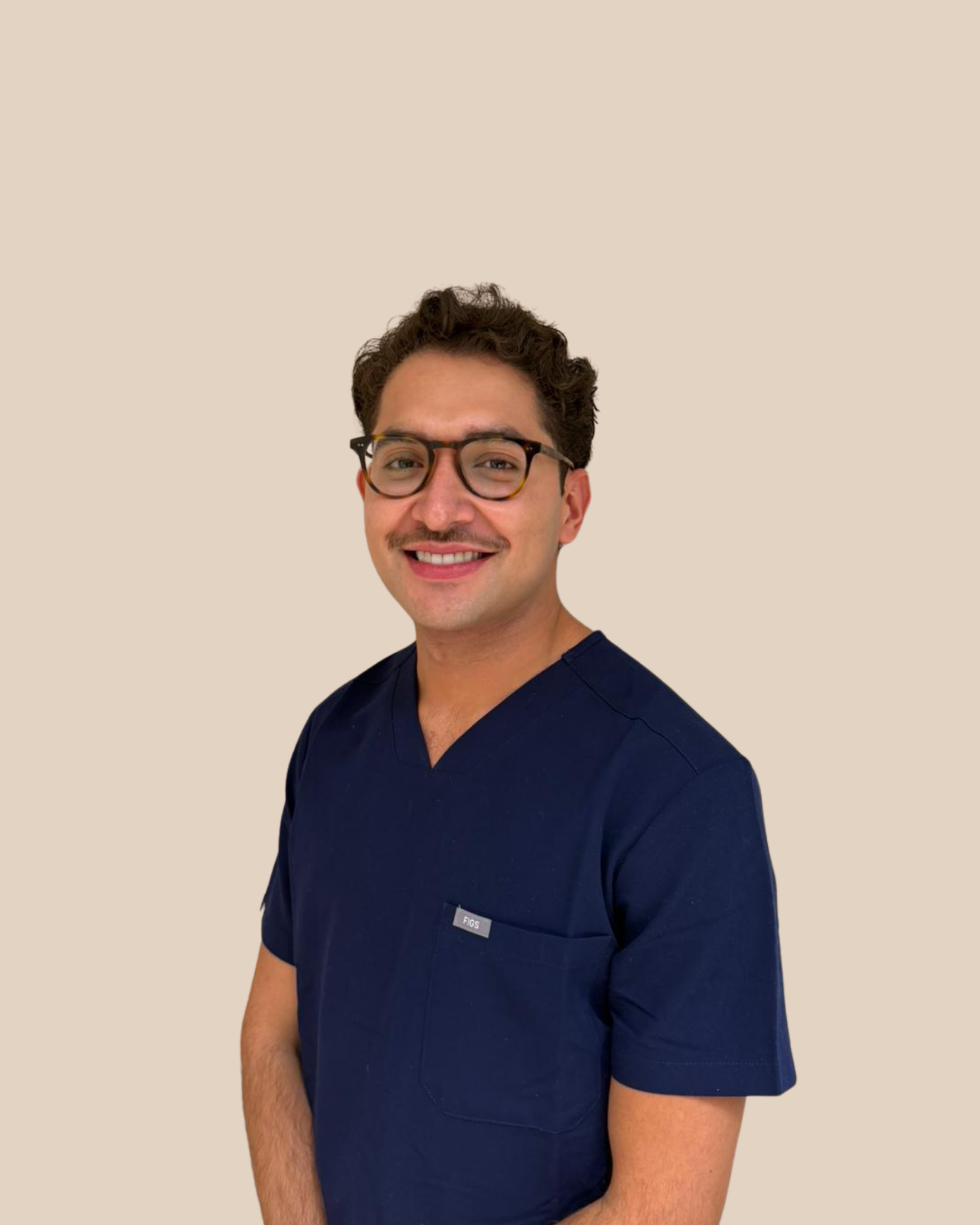Hombre con gafas y barba, vestido con uniforme médico azul, sonriendo, fondo beige.