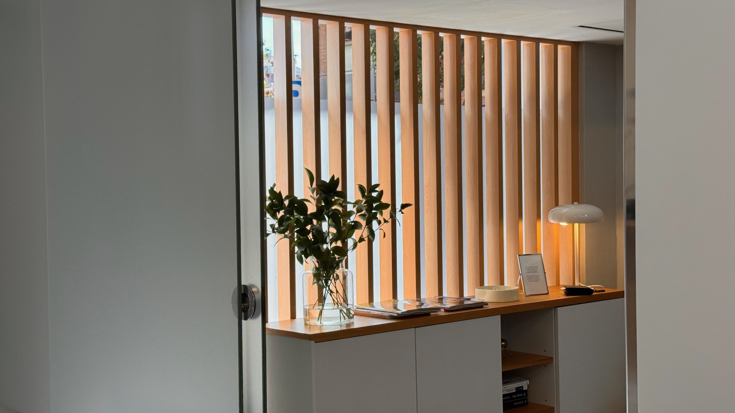 Mostrador de madera con plantas y objetos decorativos, detrás una pared con listones de madera y una lámpara blanca