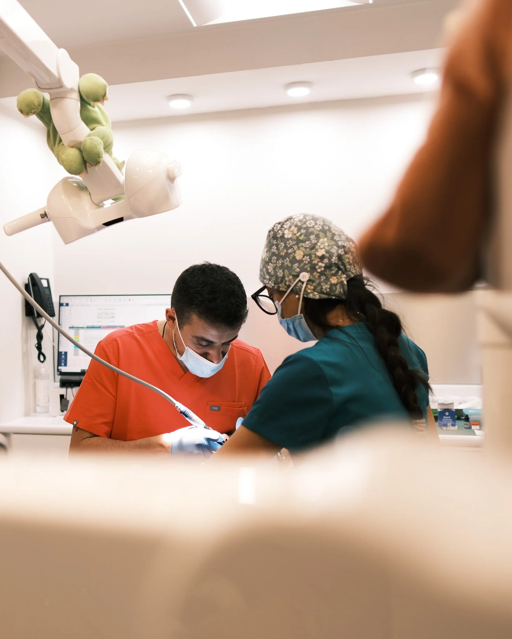 Dos profesionales de la salud, un hombre y una mujer, realizando un procedimiento médico en un consultorio, con equipo médico y monitor en el fondo.