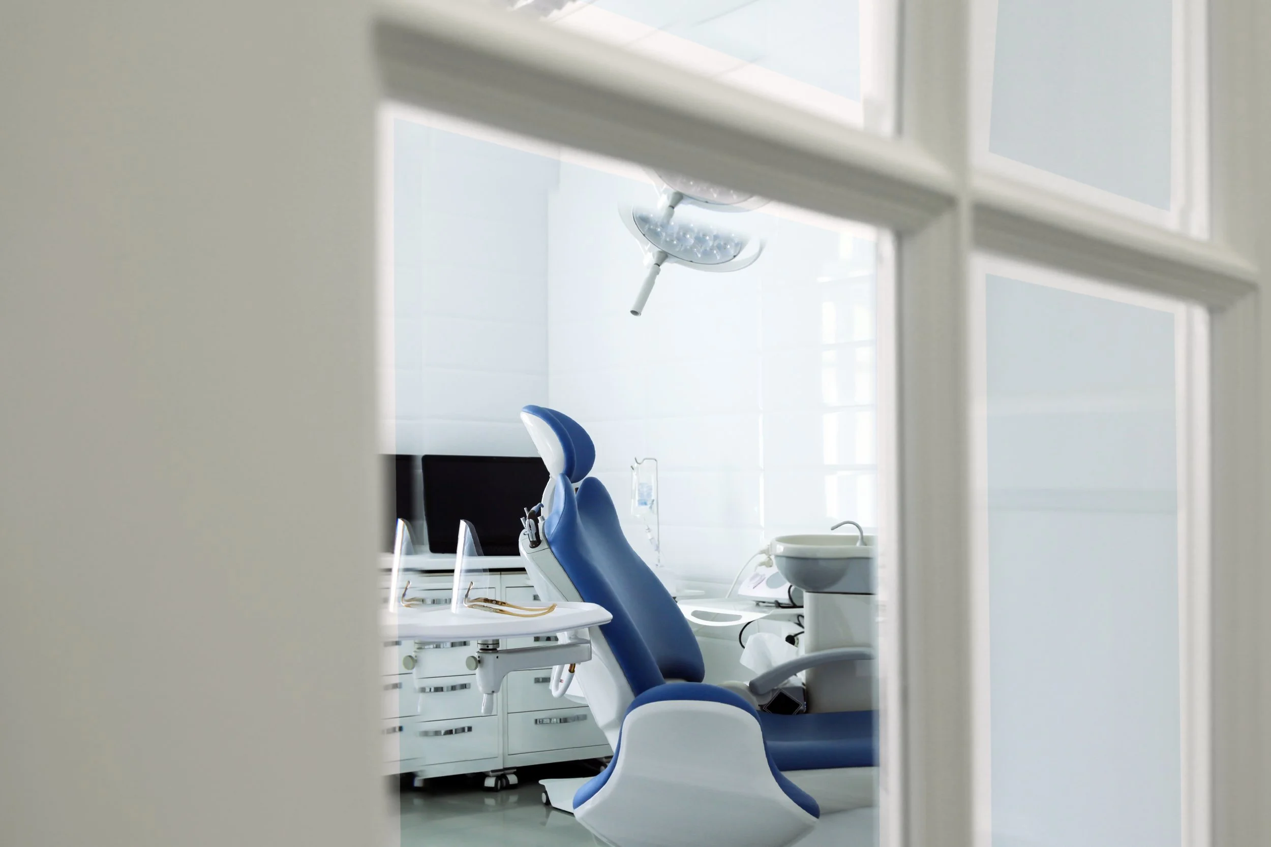 Sala de tratamiento dental con sillón azul y equipo dental, vista a través de una ventana