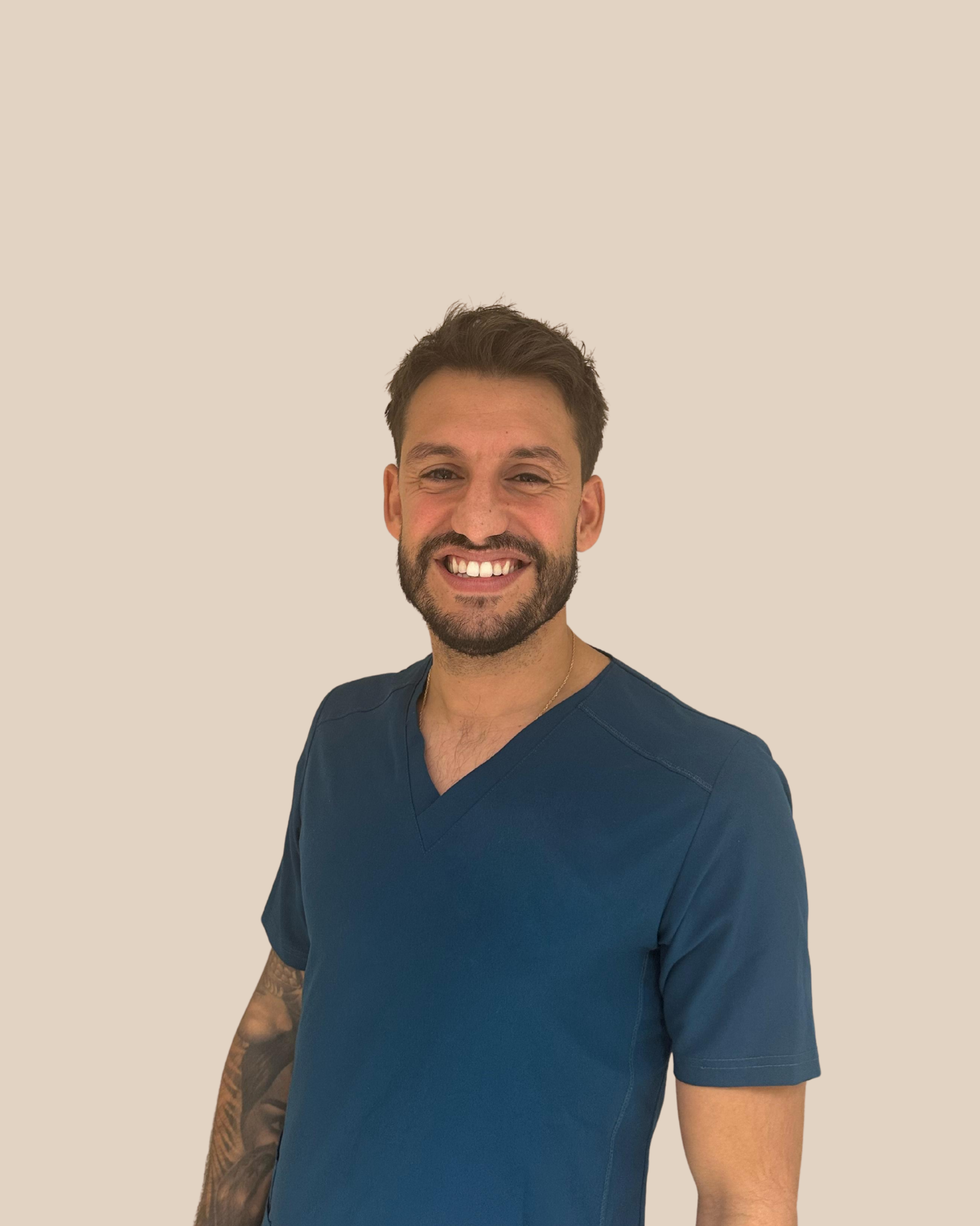 Joven hombre con barba y tatuaje en el brazo, usando uniforme médico azul, sonriendo hacia la cámara, fondo beige.
