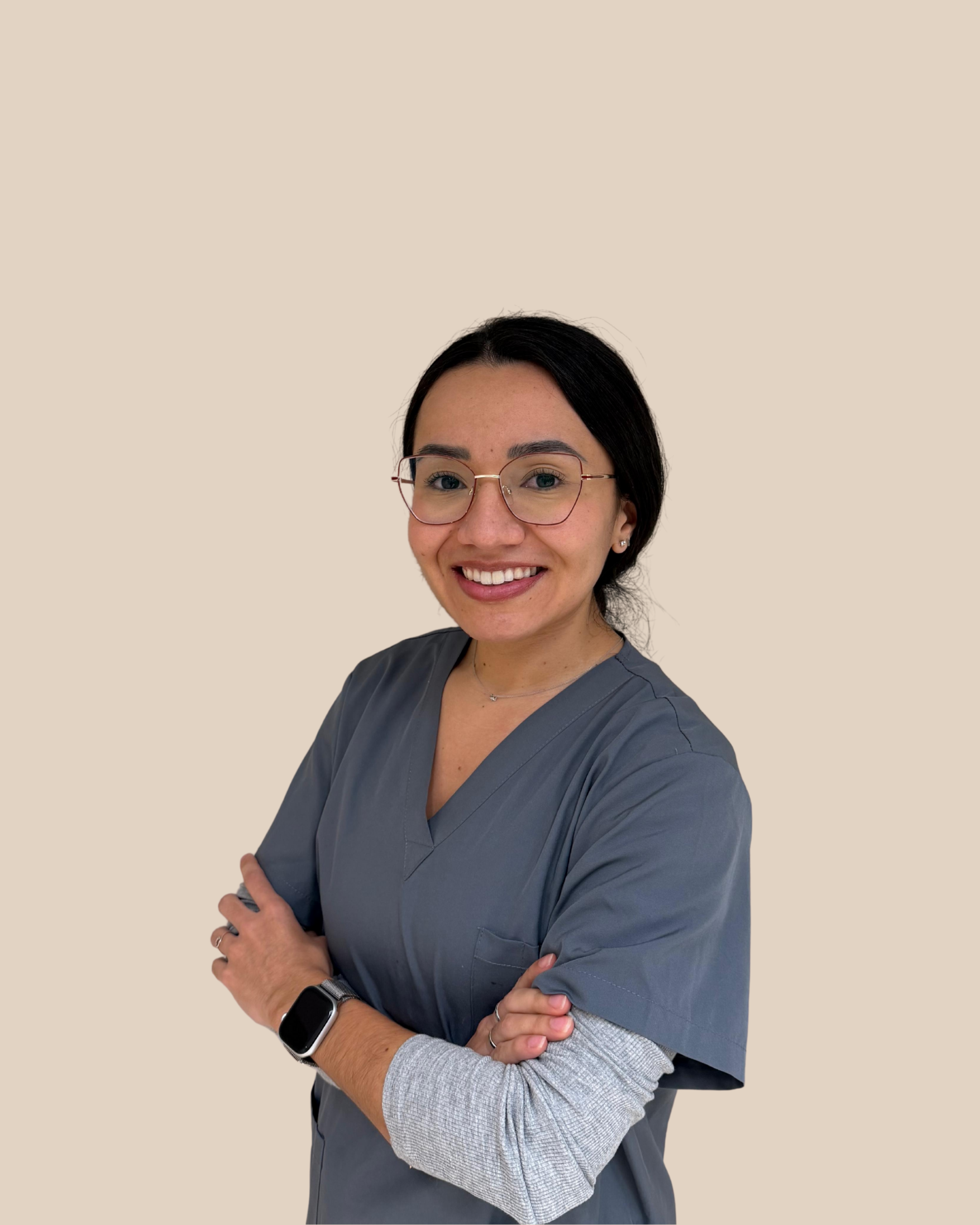 Mujer con gafas, cabello oscuro, sonriendo, con uniforme gris y fondo beige.