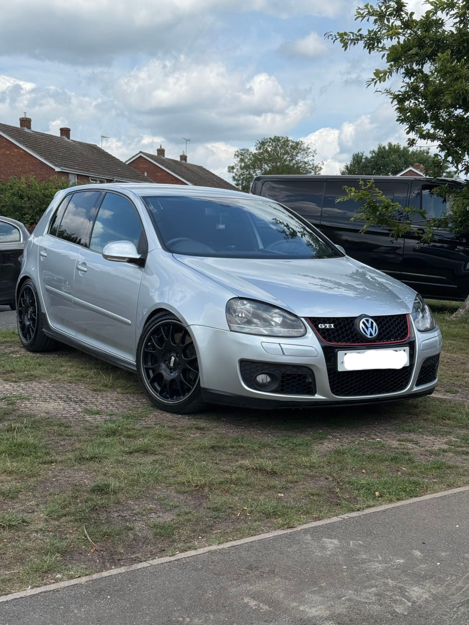 Silver Volkswagen Golf GTI parked on grass next to a sidewalk, with a black van and houses in the background under a partly cloudy sky.