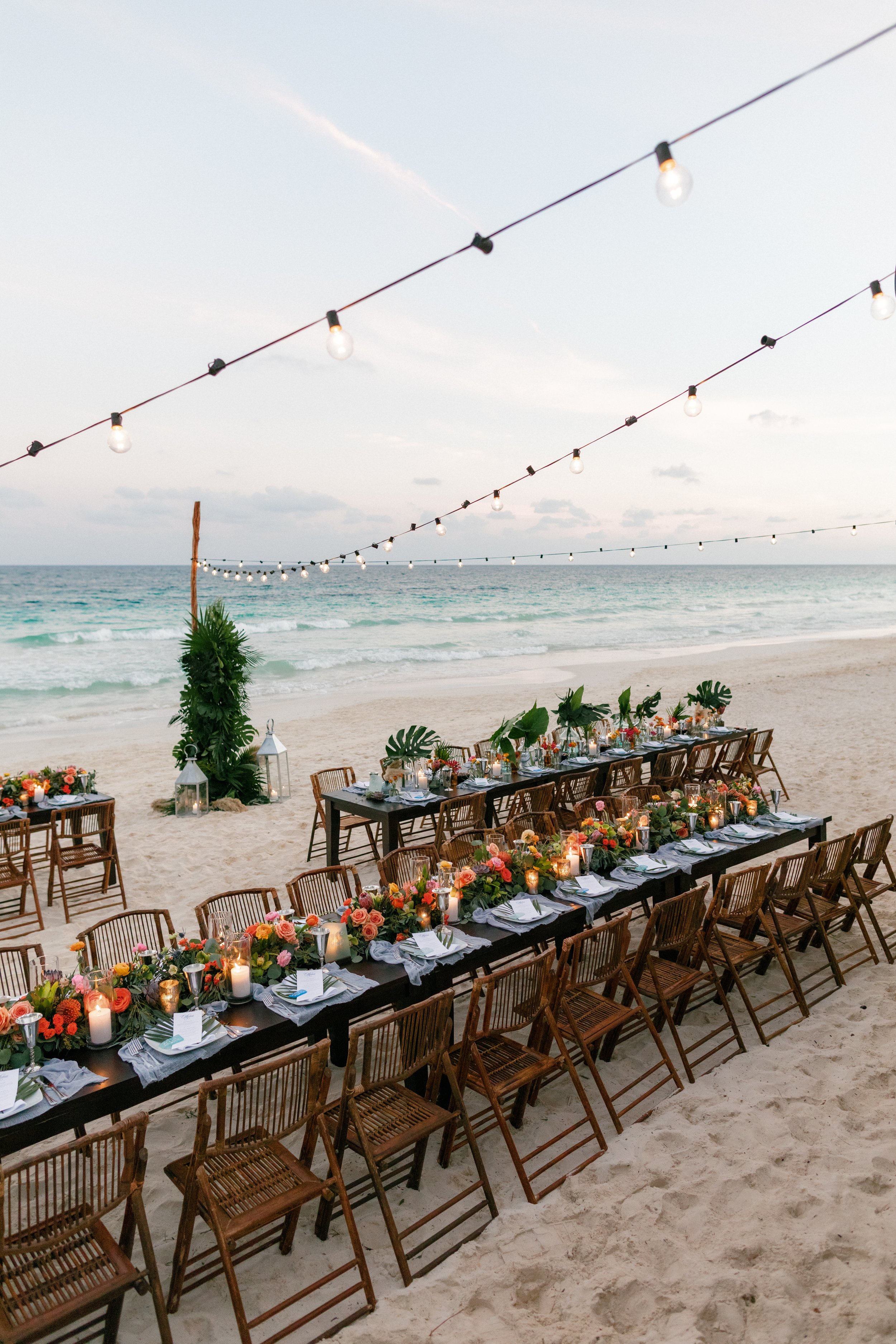 A beautifully decorated beachside dinner setup with a long table surrounded by wooden chairs, adorned with flowers in Tulum, Mexico.