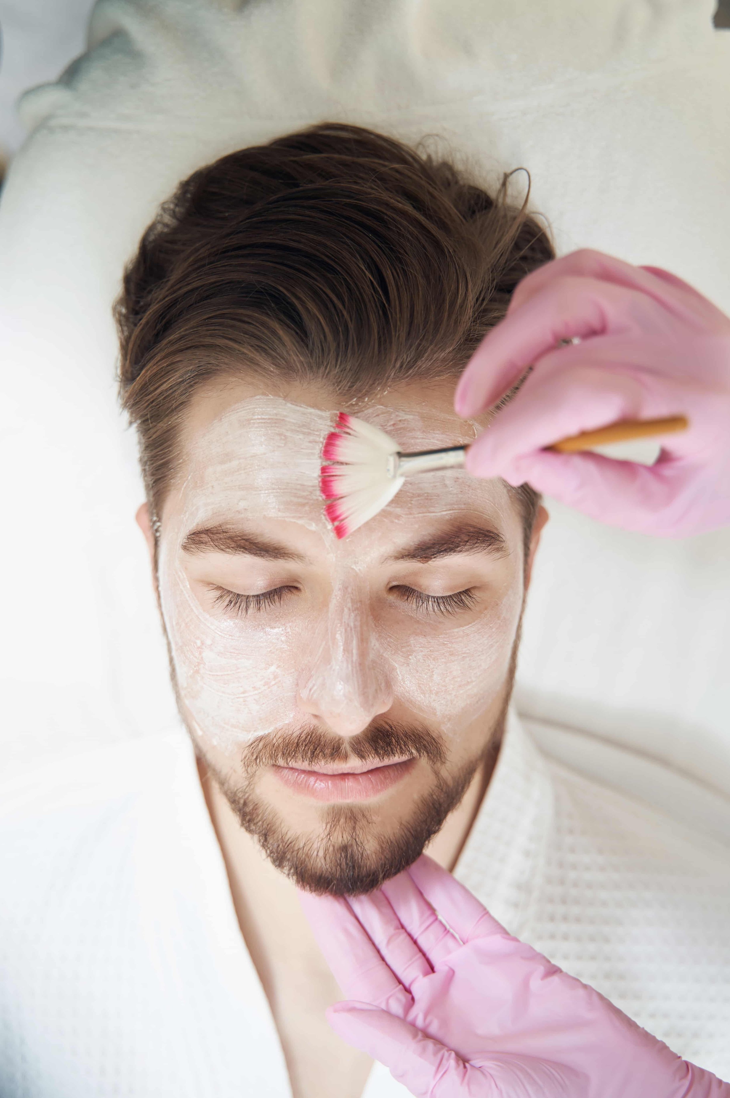 En mann får ansiktsbehandling med krem og pensel, med lukkede øyne, i en klinikk eller spa.