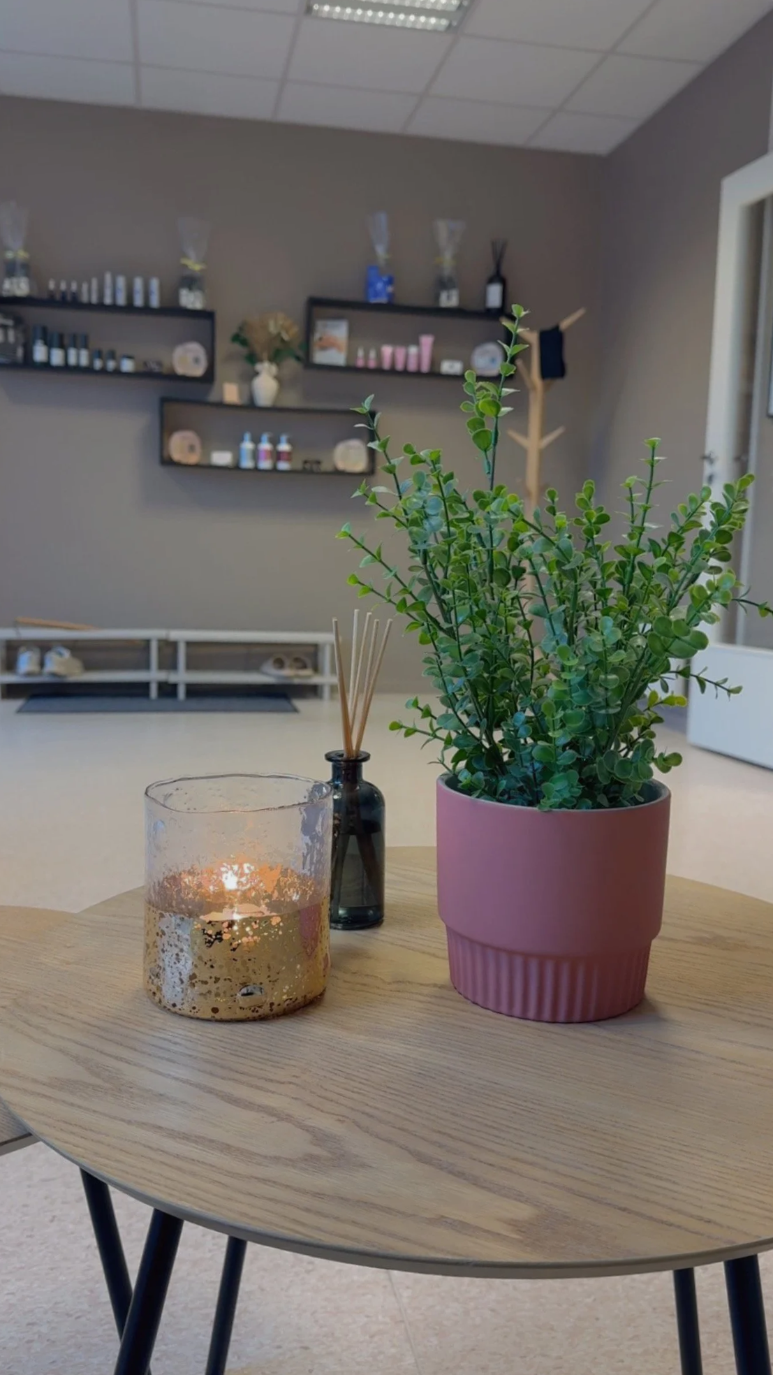 Et rundt trebord med en grønn plante i en rosa potte, et lys i en gullfarget holder og en liten svart glassflaske med teskjeer inni.