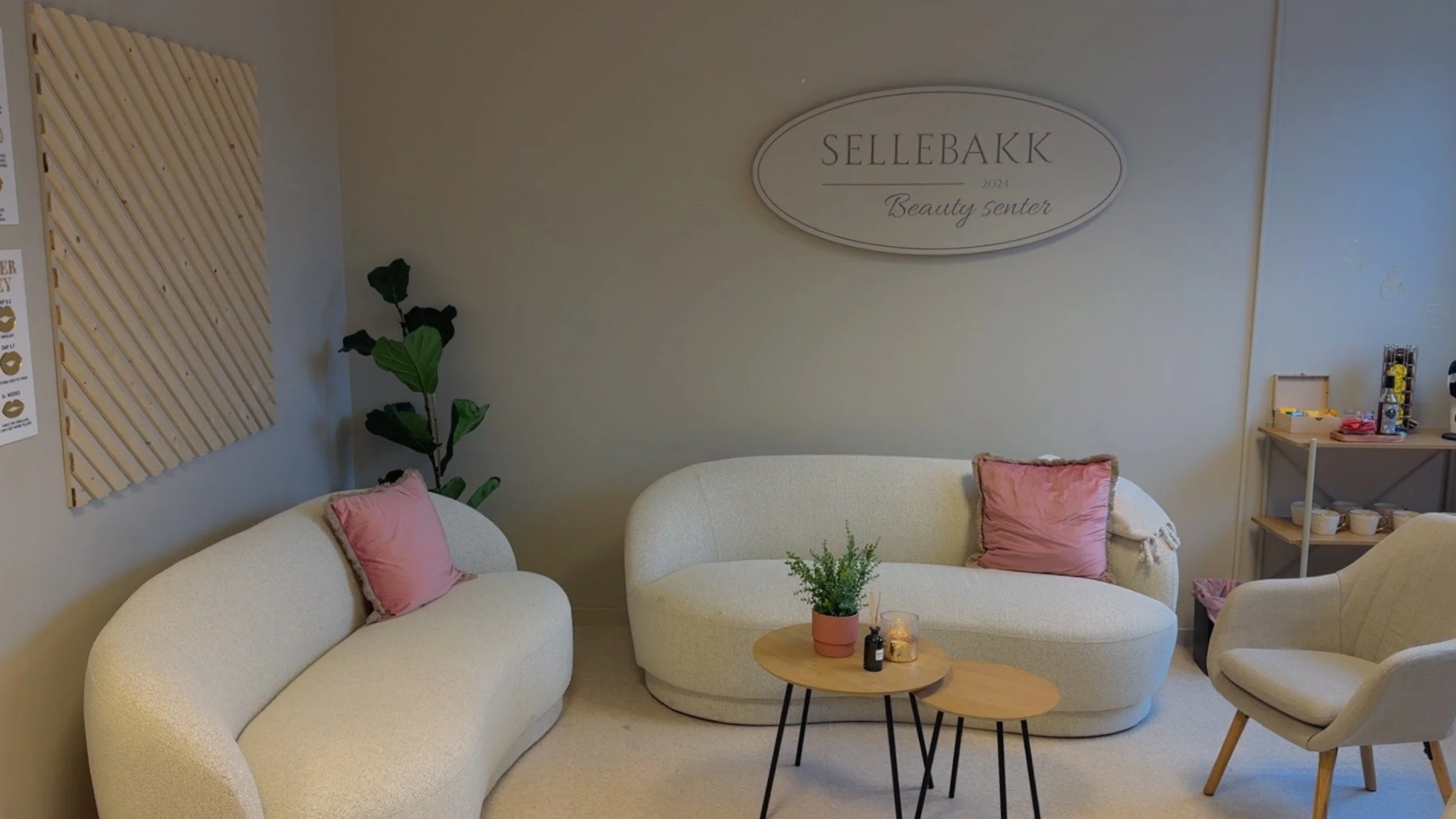 Loungemøteområde med hvite sofaer, rosa puter, et lite rundt bord med planter og lys, og et merke som sier Sellebakk 2024 Beauty senter. Det er en grønn plante i hjørnet og en hyller med diverse gjenstander på høyre side.