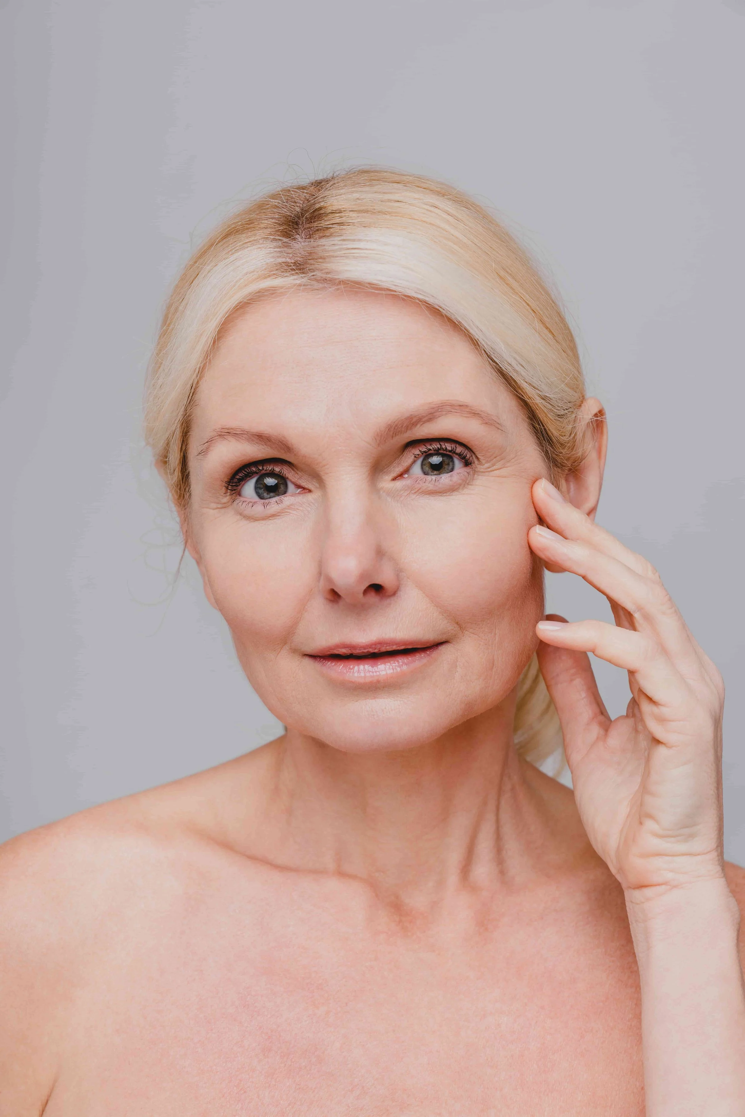 En eldre kvinne med blondt hår og blå øyne, ser rett mot kamera og berører sitt kinn.