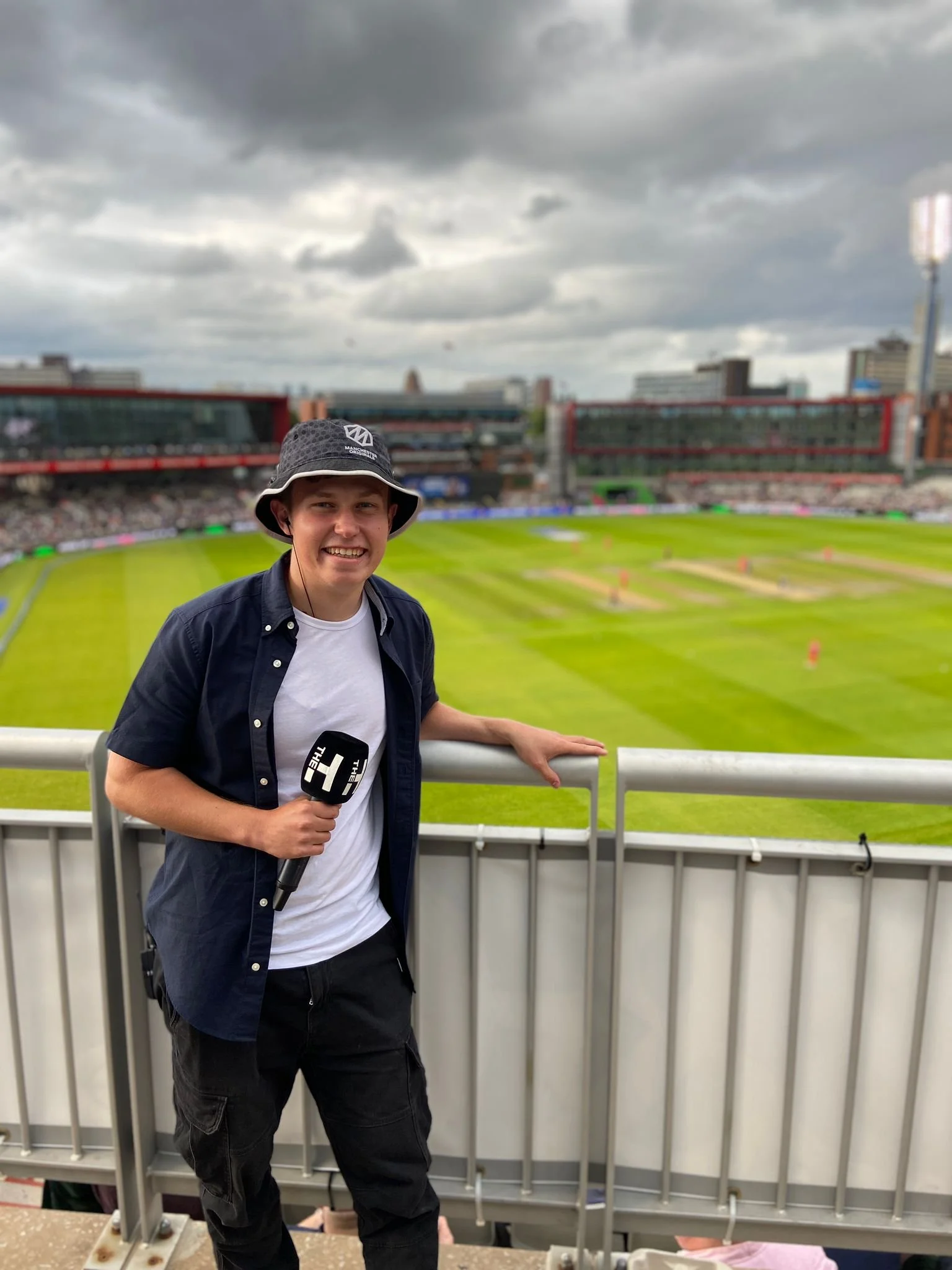 Simon Reynolds the sports presenter holding a microphone at a cricket stadium with a lush green field and cloudy sky in the background.