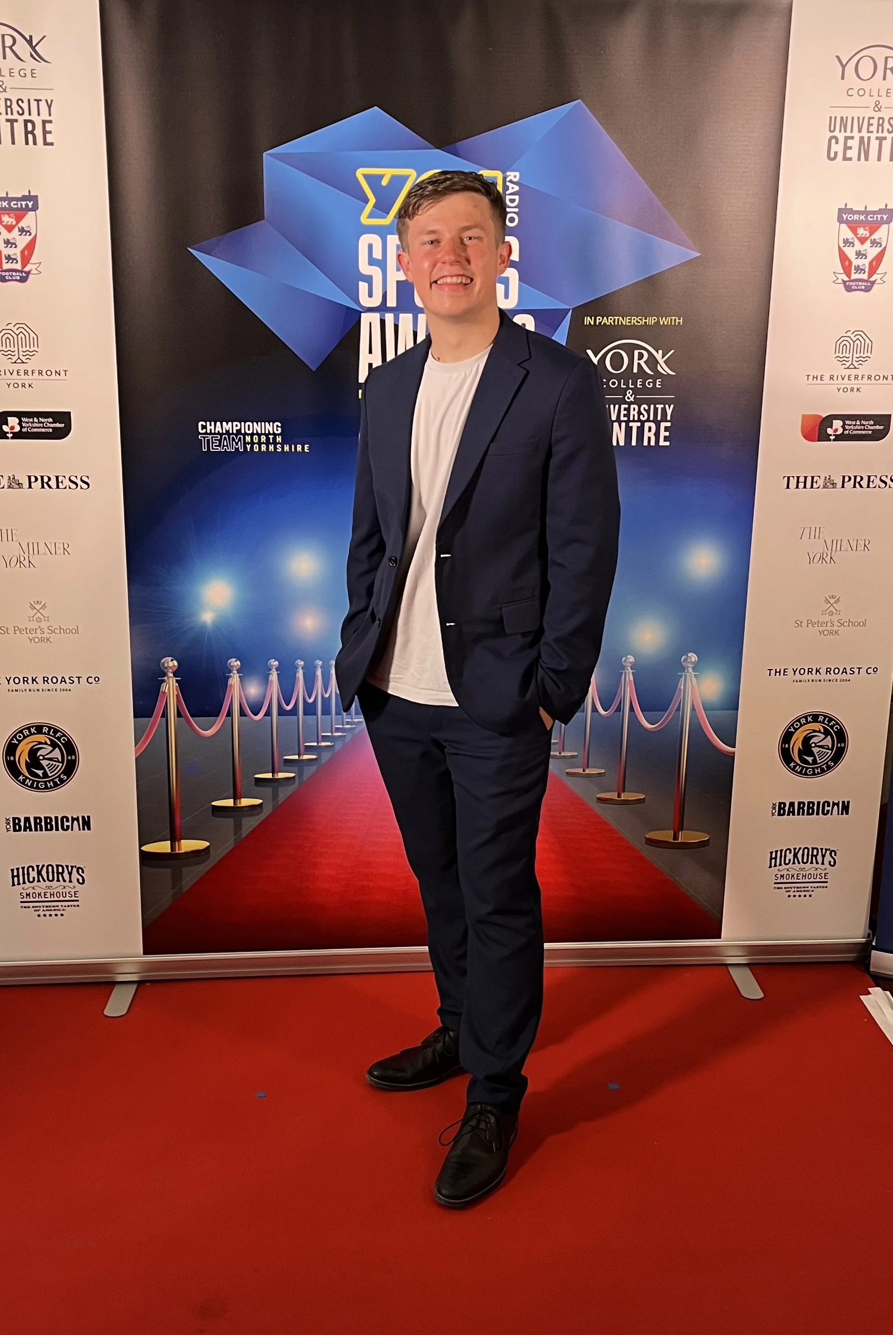 Presenter and Sports Host in a navy suit and white t-shirt standing on a red carpet with a backdrop displaying logos and event branding for a sports awards event.