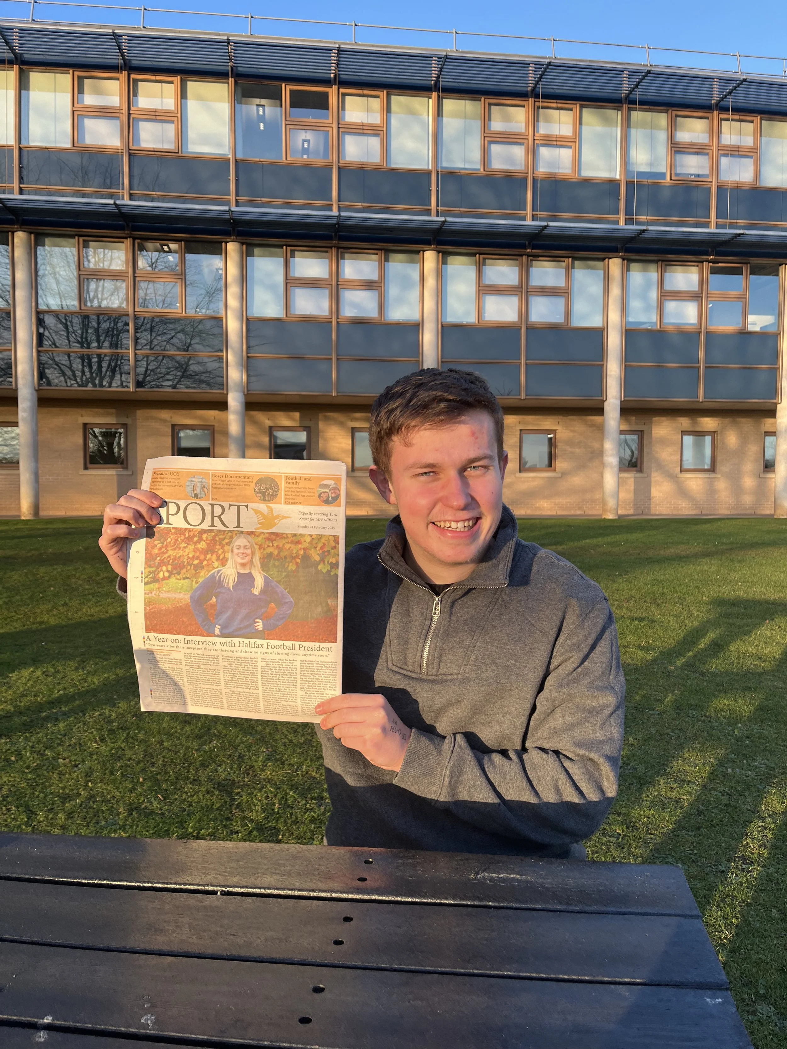 A young man sitting at a park bench outdoors, holding up a newspaper with a smiling face. Behind him is a modern building with large reflective windows and a blue sky with some clouds.