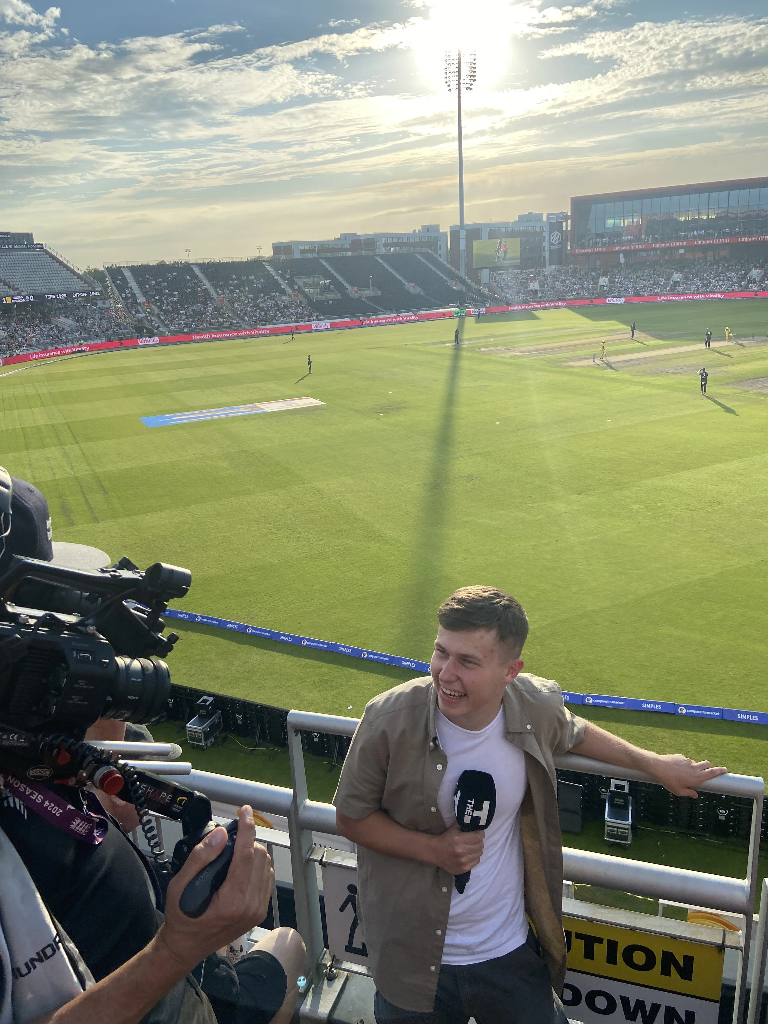 Simon Reynolds standing at the edge of a cricket stadium holding a microphone, being filmed or interviewed by a cameraman, with a green field and spectators in the stands in the background. Hosting the cricket and doing sports presentation.