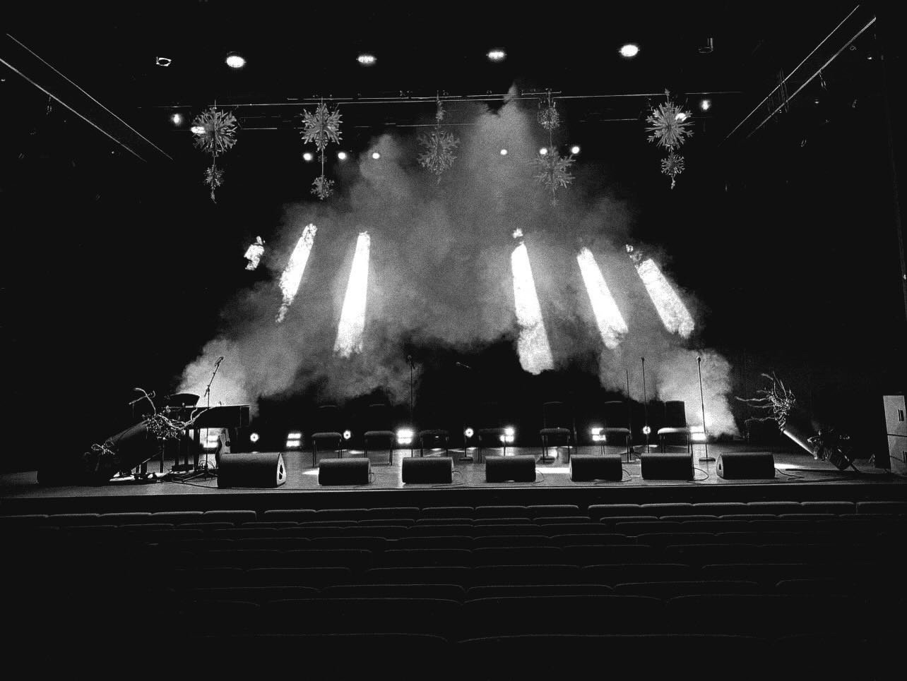 Empty theater stage with fog, bright lights, snowflake decorations hanging from the ceiling, and microphones on stand, preparing for a performance.