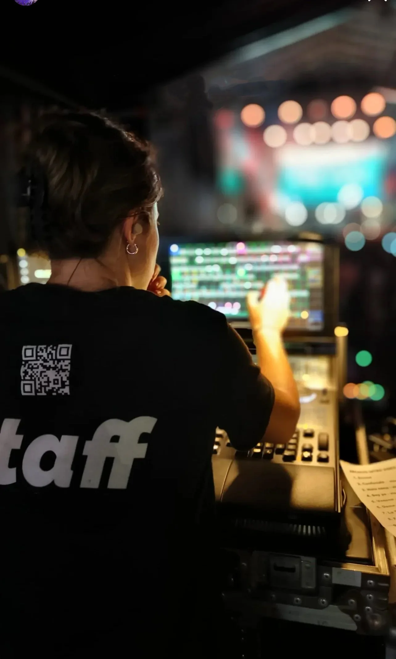 A DJ stands at a mixing console with a laptop, playing music at a live event or party, with colorful stage lights in the background.