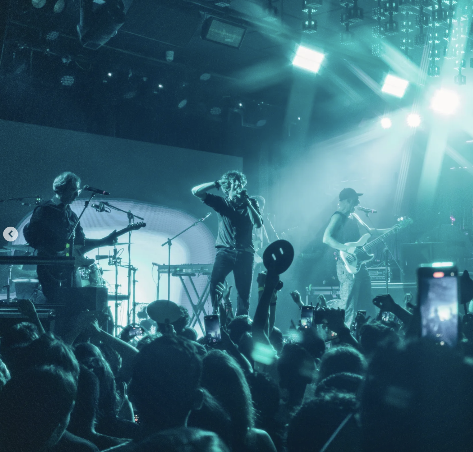 A band performing live on stage with bright stage lighting, a crowd in the foreground, some audience members holding phones, and the lead singer in the center with curly hair, wearing dark clothing and holding a microphone.