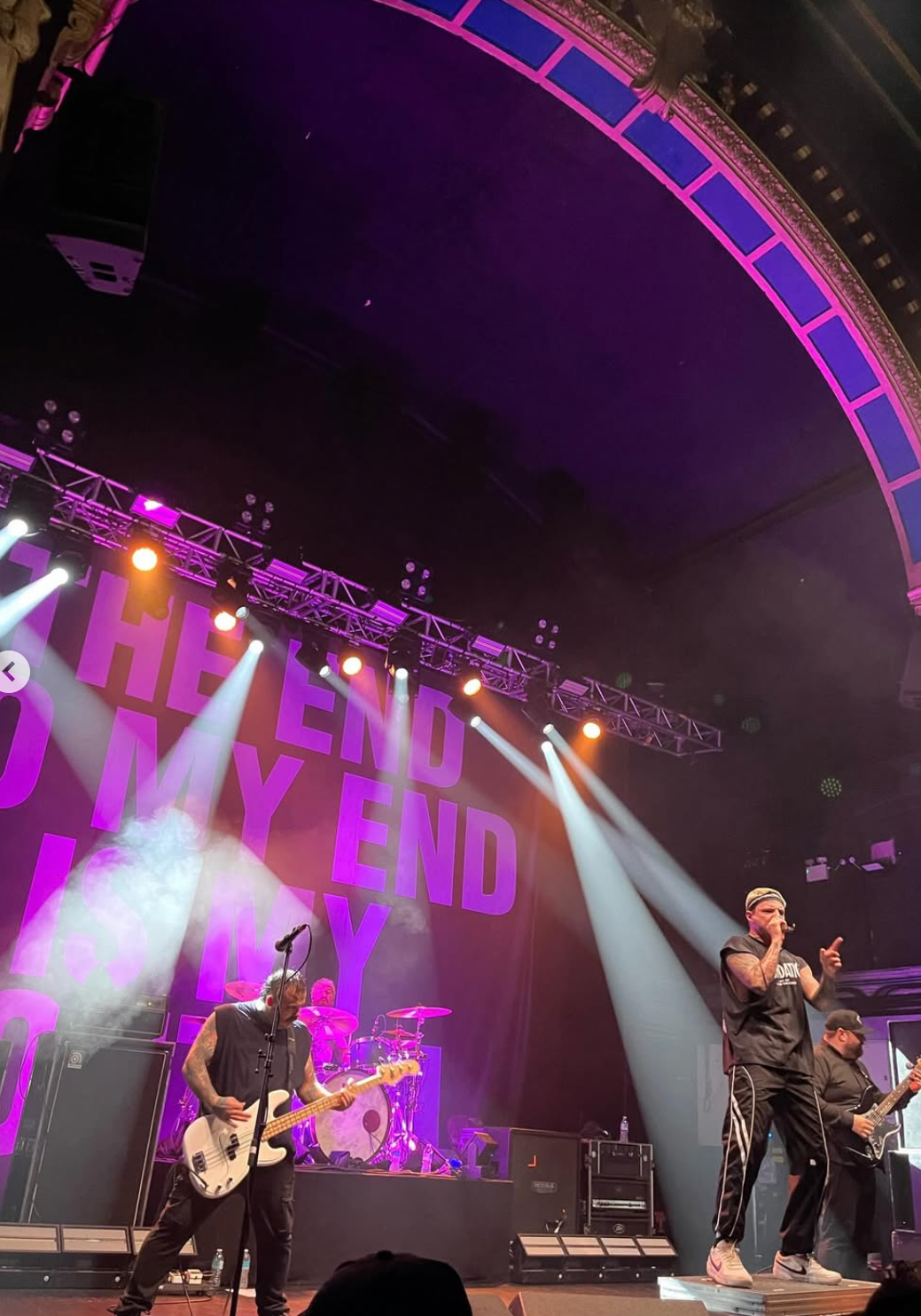 Musicians perform on stage with colorful lighting and a backdrop displaying the words "THE END MY END," with the lead singer holding a microphone and wearing sunglasses, while other band members play guitars and drums.