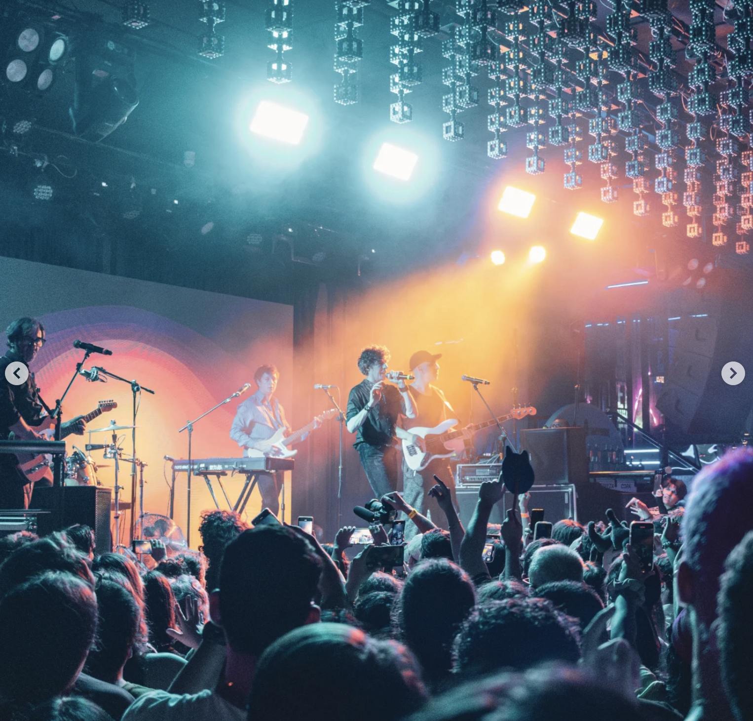 A band performs on stage at a concert with multi-colored lights overhead and a crowd of audiences in front, many capturing the moment on their phones.