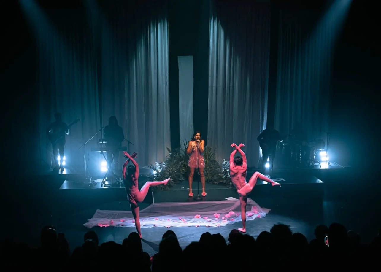 Stage performance with a singer in the center, two ballet dancers in pink costumes, and a band with musicians in the background, illuminated by blue stage lights.