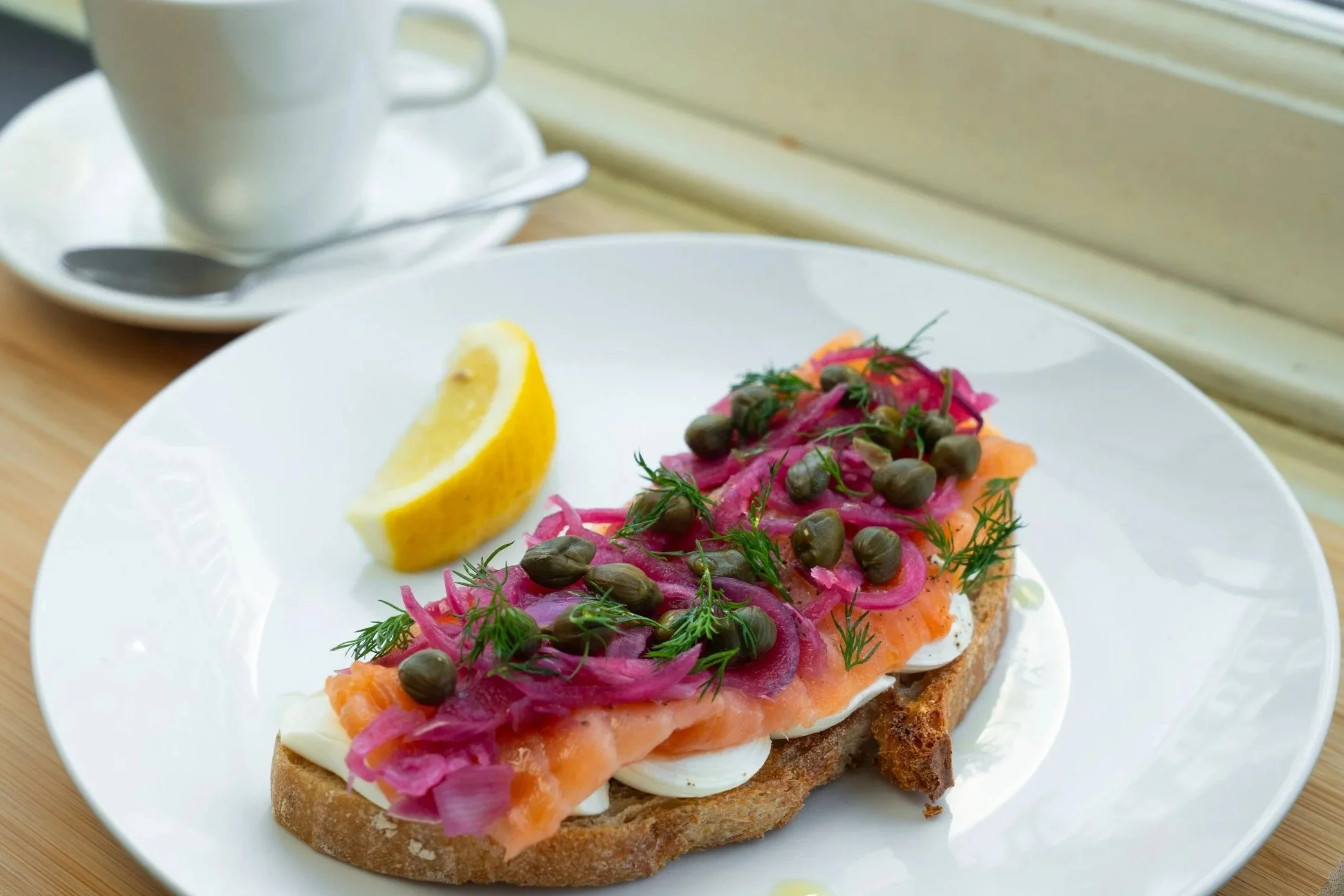 Open-faced smoked salmon sandwich with pickled onions, capers, dill, and lemon wedge on a white plate, with a white cup of coffee or tea in the background.