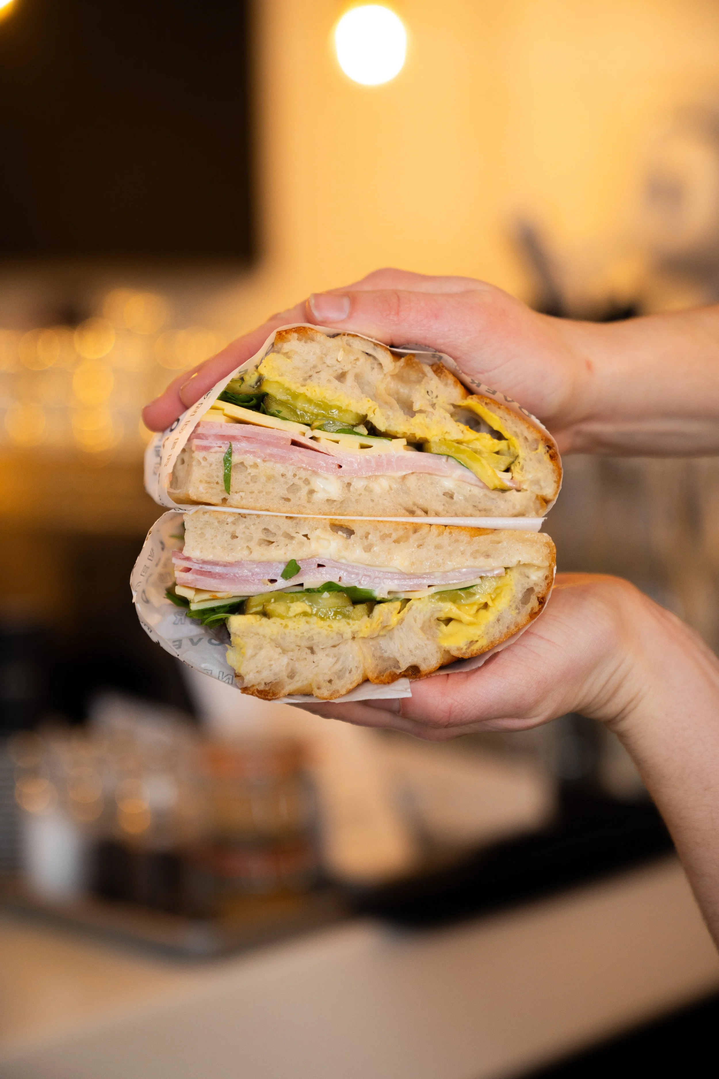 A person holding a cut sandwich with ham, cheese, cucumber, and egg, in a cafe or restaurant setting.
