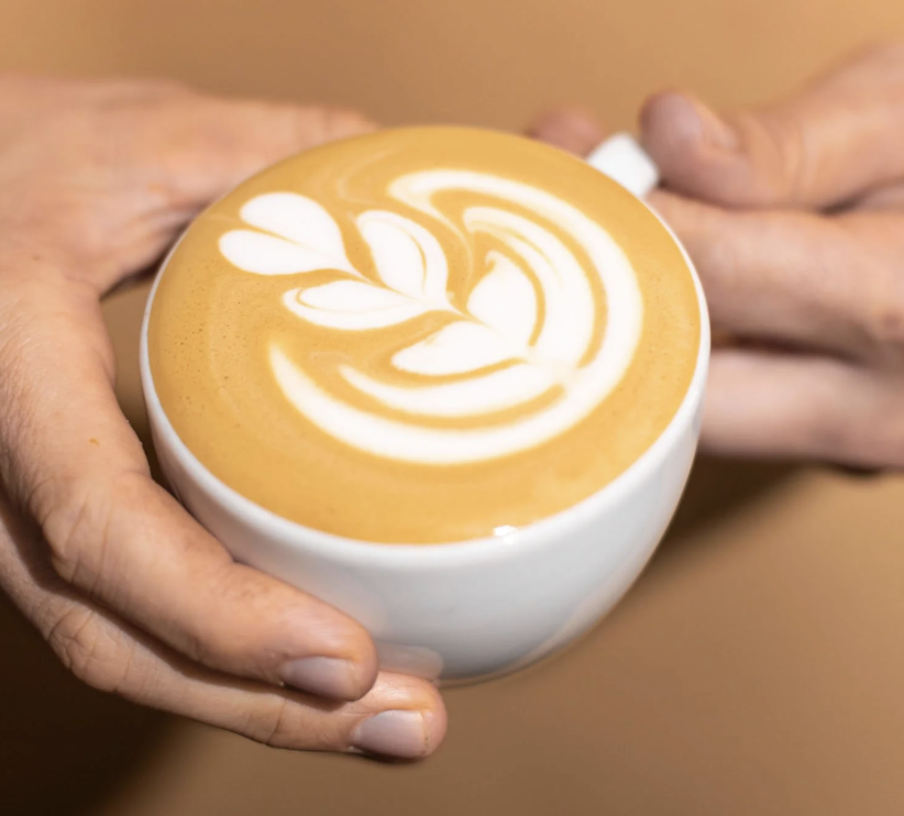 A person holding a white ceramic cup of latte with latte art in the shape of a floral pattern.
