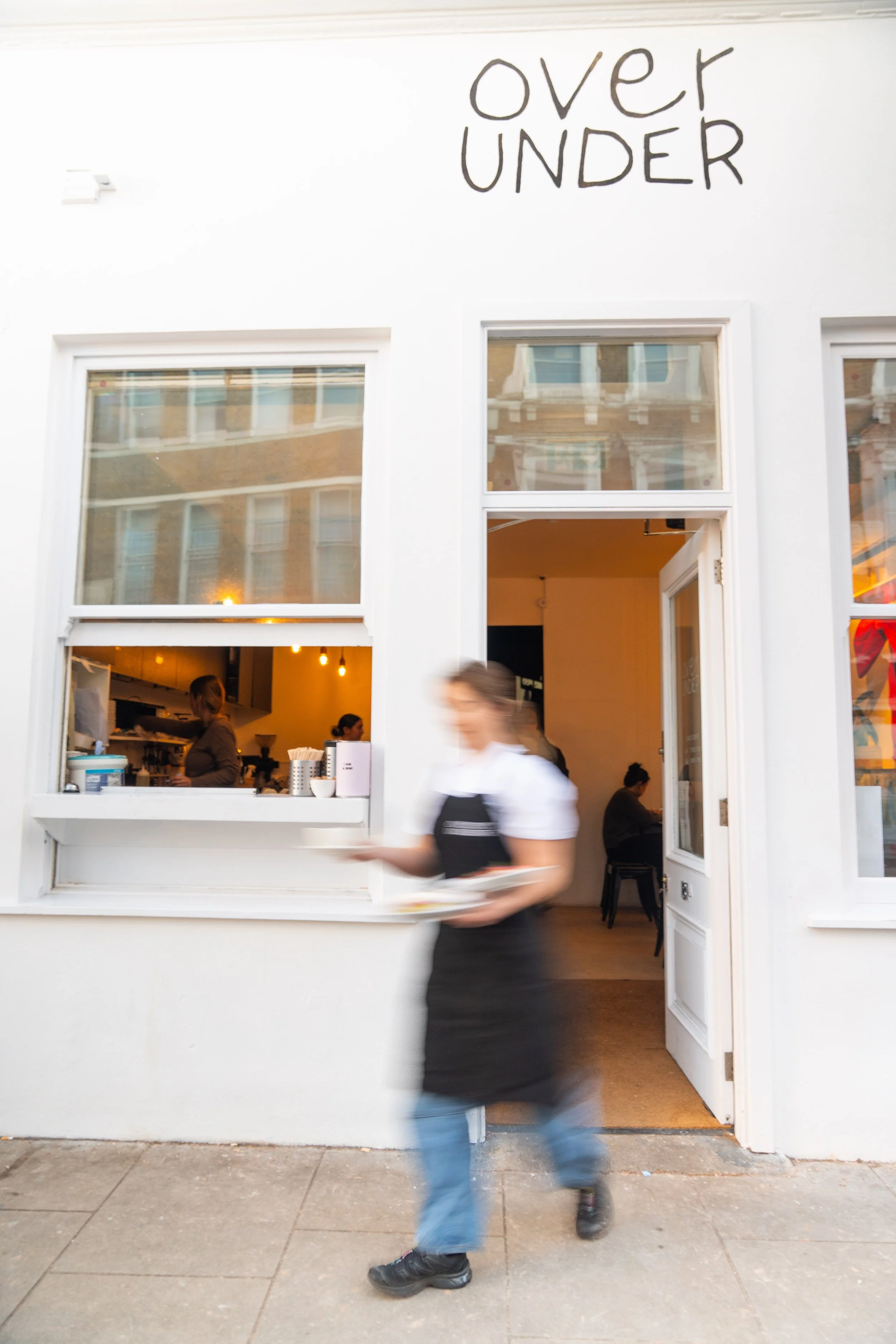 A blurred waitress wearing a black apron and blue jeans serving a dish outside a white restaurant with a sign that says "over" on top and "UNDER" below, with visible interior through open window and door.