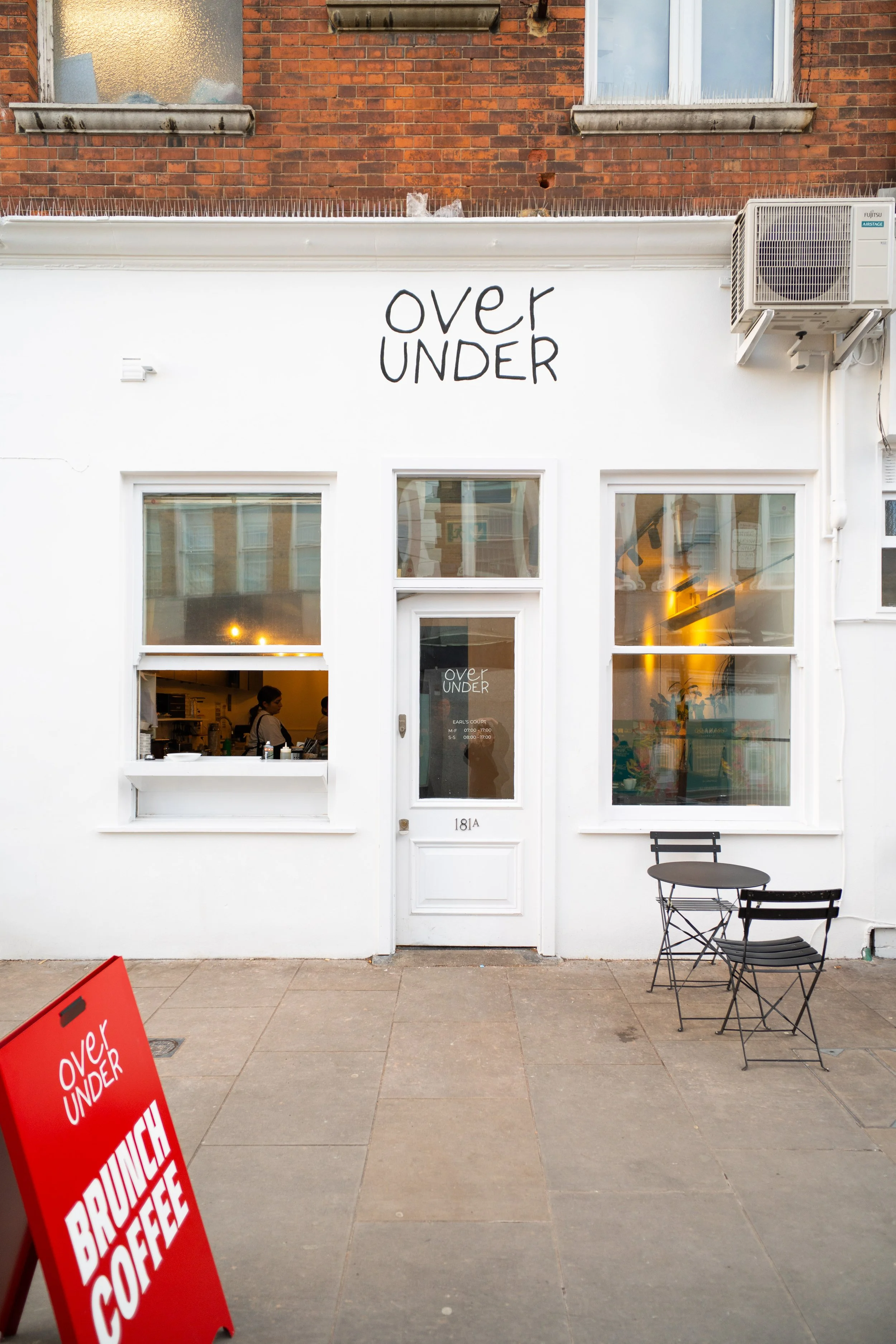White storefront with black handwritten sign "over UNDER" above the door. Two large windows reveal a cozy interior with warm lighting and a person inside. A small outdoor table with two chairs is placed outside, and a red sandwich board sign with the same "over UNDER" logo advertises brunch and coffee. An air conditioning unit is mounted on the upper right corner of the building.