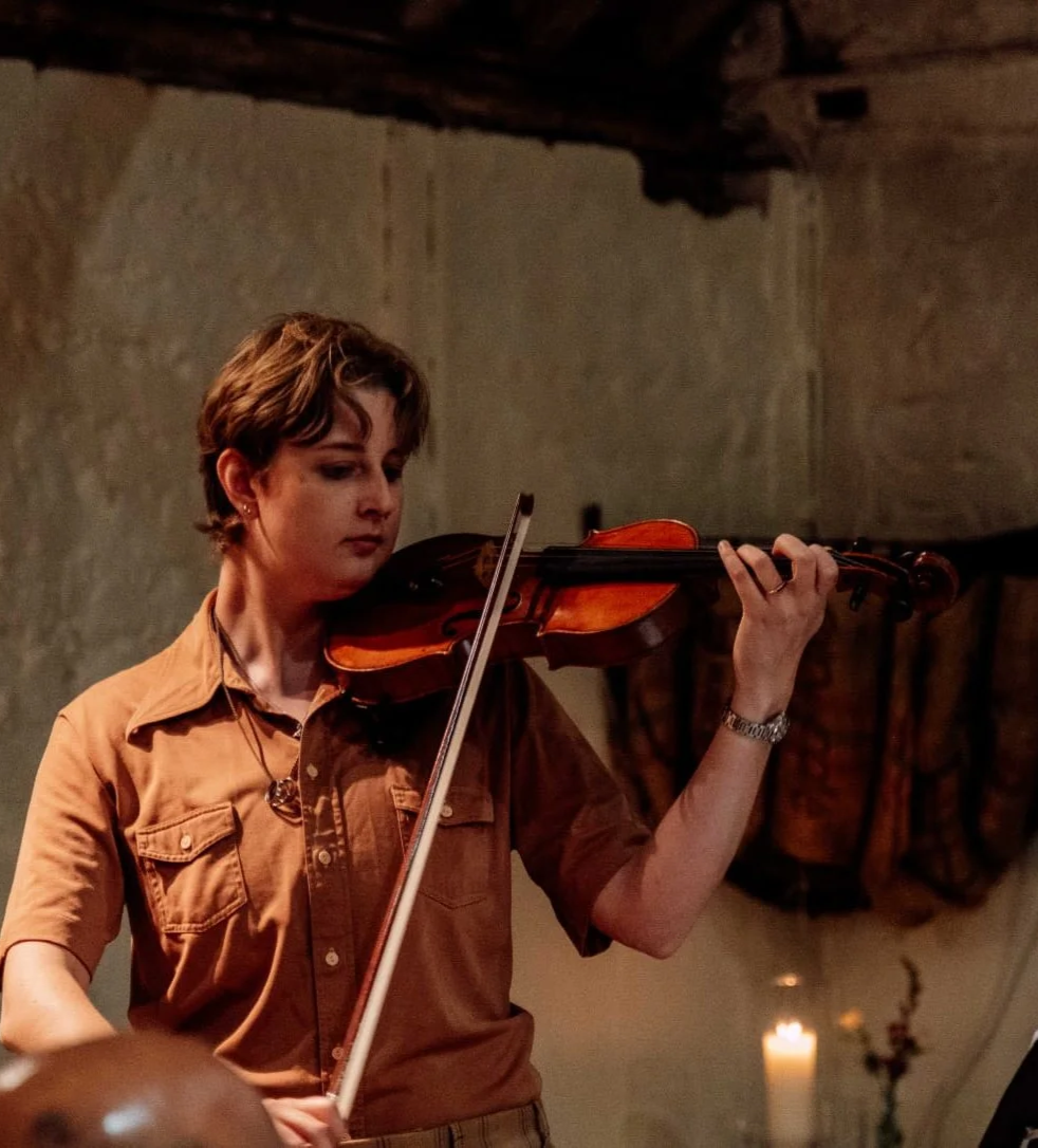 A woman playing the violin indoors, with candles and other objects in the background.