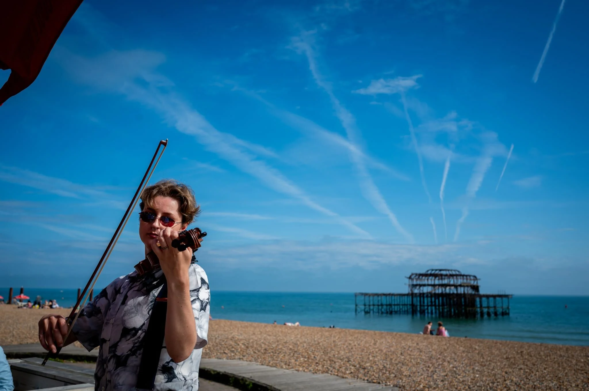 A person with short hair, sunglasses, and a patterned shirt playing a violin on a beach with a peeled pier in the background and a bright blue sky with contrails.