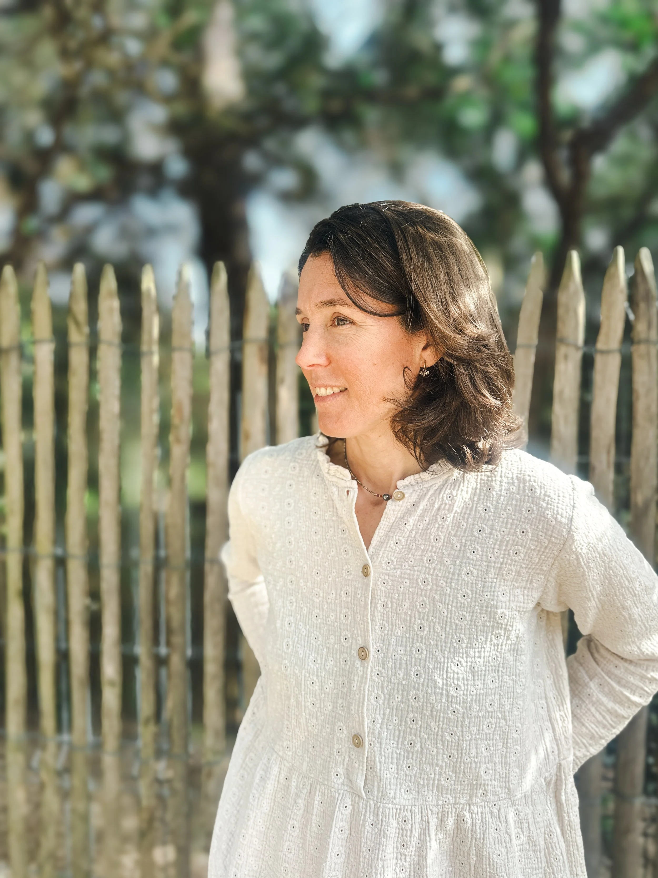 Laure praticienne Monbourquette spécialisée estime de soi accompagnement individuel. Femme souriante portant une robe blanche avec un fond de clôture en bois et des arbres en arrière-plan.Dune du Pilat. Gironde. Bassin d'arcachon. Bordeaux