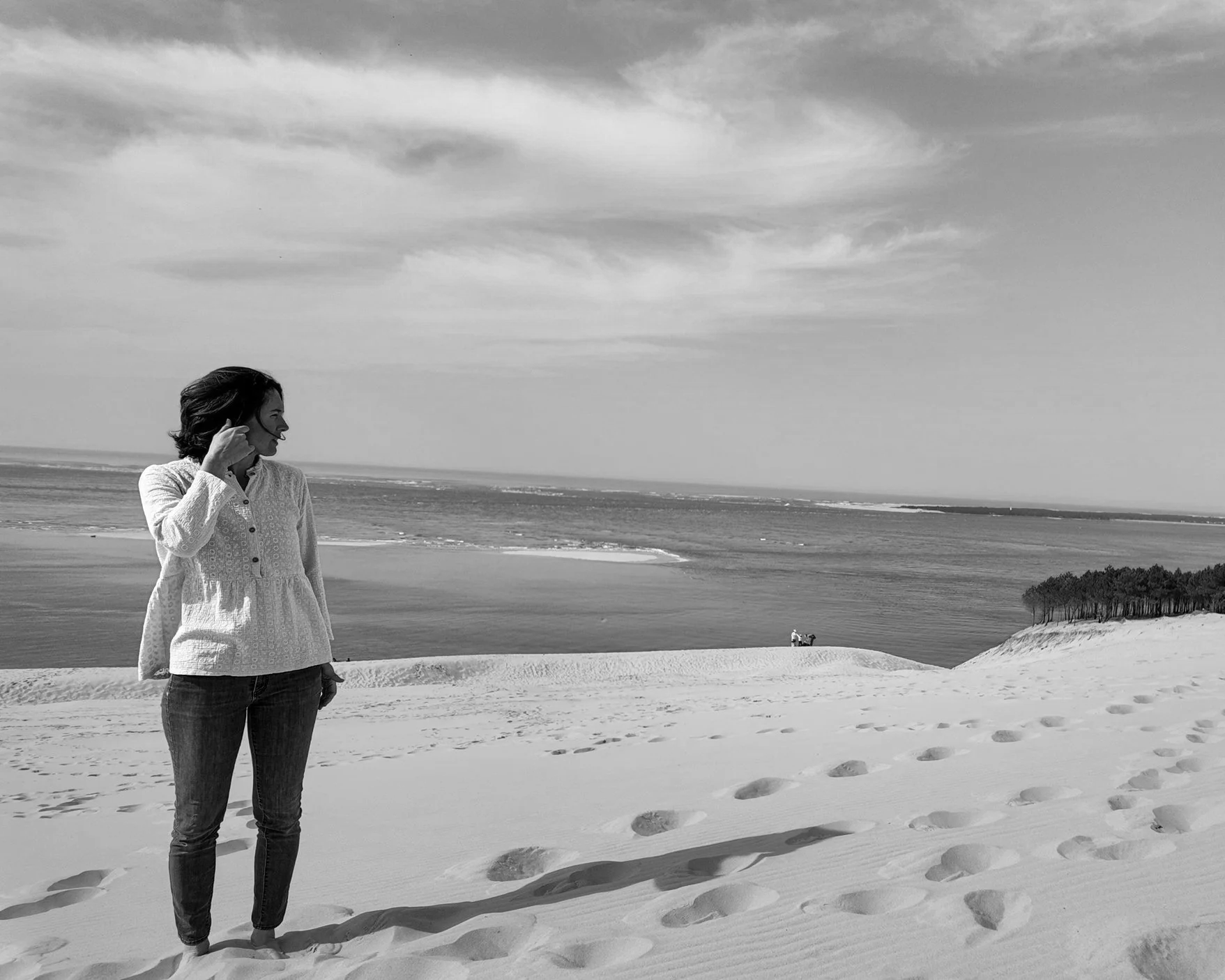 Séance estime de soi méthode Monbourquette coaching développement personnel. Une femme debout sur une plage de sable, regardant vers la gauche, avec l'océan en arrière-plan, des dunes de sable et un groupe d'arbres au loin. Dune du Pilat bordeaux