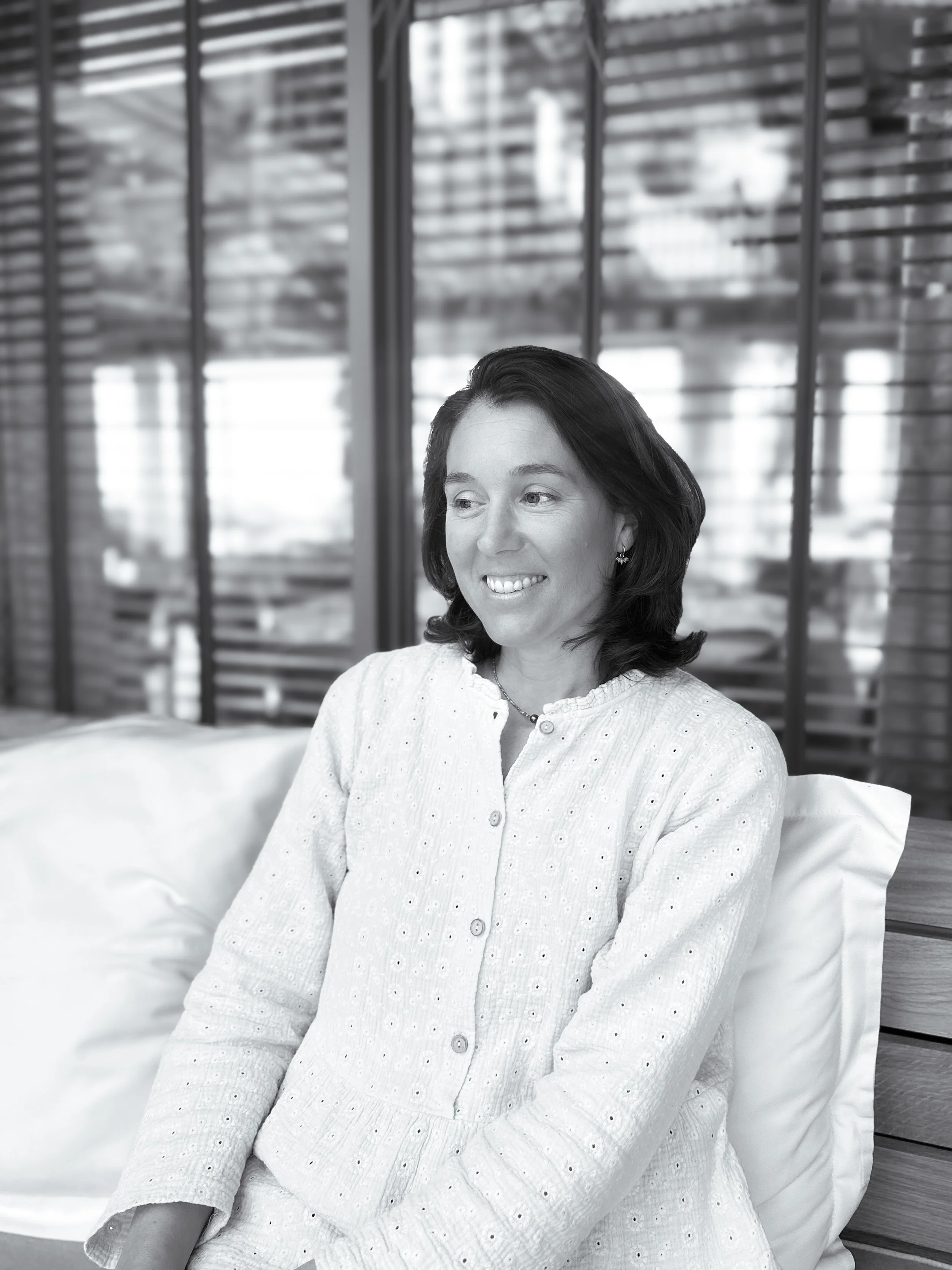 "Laure praticienne estime de soi méthode Monbourquette consultation Bassin d’Arcachon. Une femme souriante assise sur un canapé avec un fond de grands volets en bois et un bâtiment en verre derrière.