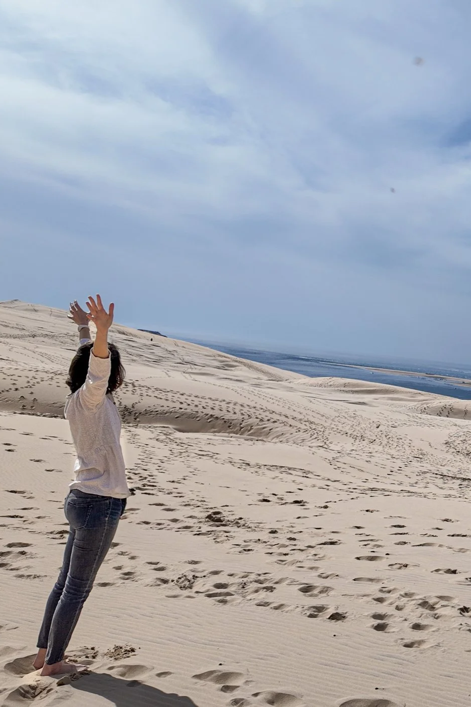 Libération émotionnelle méthode Monbourquette confiance en soi accompagnement. Une femme debout dans un désert de dunes de sable, levant les bras vers le ciel, avec un ciel nuageux au-dessus et une étendue d'eau au loin. Gironde. Bordeaux