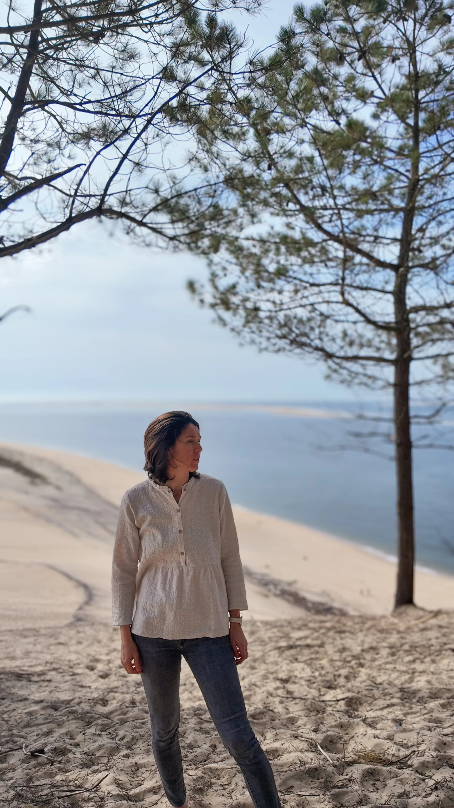 "Praticienne estime de soi méthode Monbourquette accompagnement développement personnel.Une femme debout sur une plage de sable, sous un arbre, avec la mer en arrière-plan, un ciel nuageux. Dune du Pilat