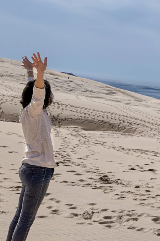 Femme debout sur une plage de sable, levant les bras, avec des dunes de sable et la mer en arrière-plan.