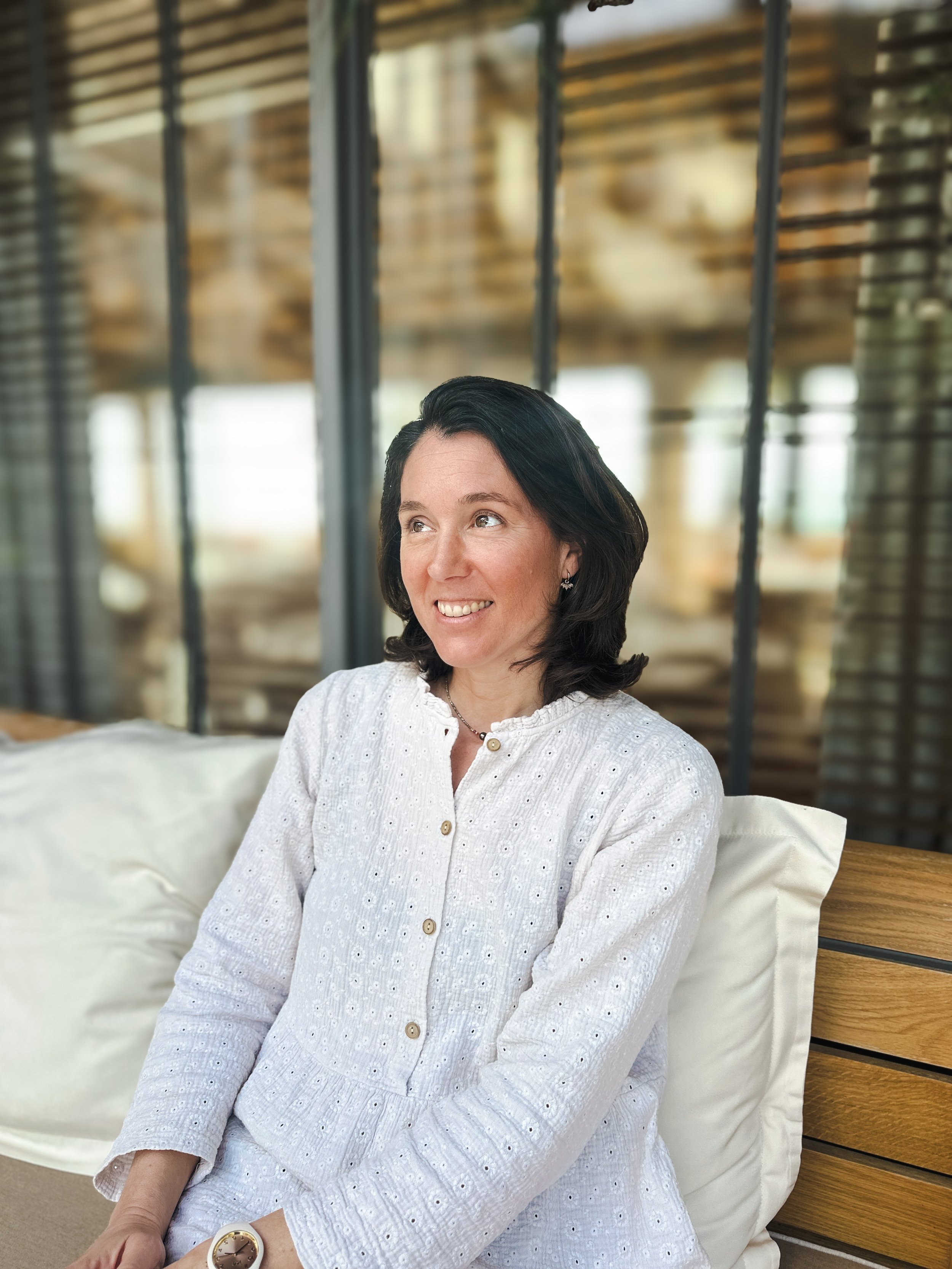 Accompagnement confiance en soi praticienne Monbourquette consultation bien-êtreUne femme aux cheveux noirs, souriante, portant une robe blanche en maille, assise sur un canapé en bois et en tissu blanc, dans un lieu moderne avec de grandes fenêtres