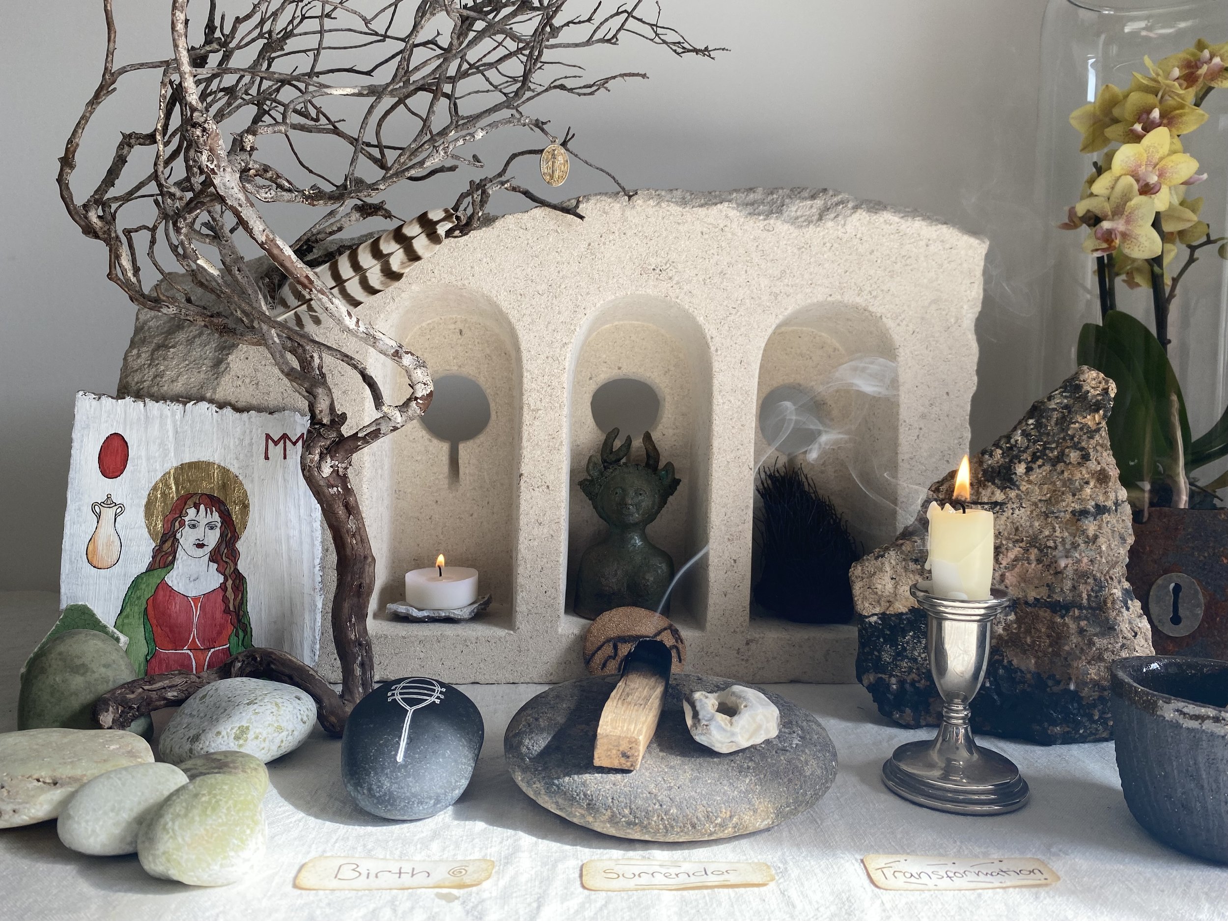 A collection of rocks, stones, and shells arranged on a surface with a painting and decorative items in the background, including a small tree branch, candles, and plants. There are labels with words like 'Birth 6,' 'Surrender,' and 'Transformation' beneath the arrangement.