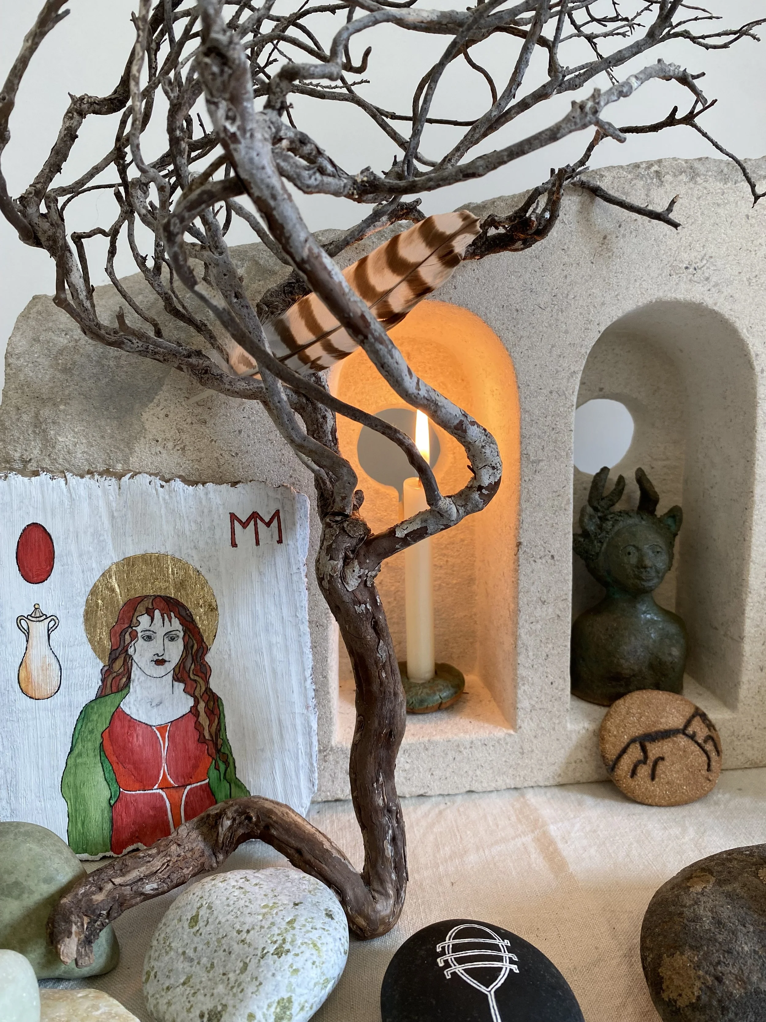 A decorative arrangement featuring a twisted tree branch in front of a lit candle inside a small alcove, with stones and a painting of a woman with red hair and green cloak, and a small statue of an animal head in a niche to the right.