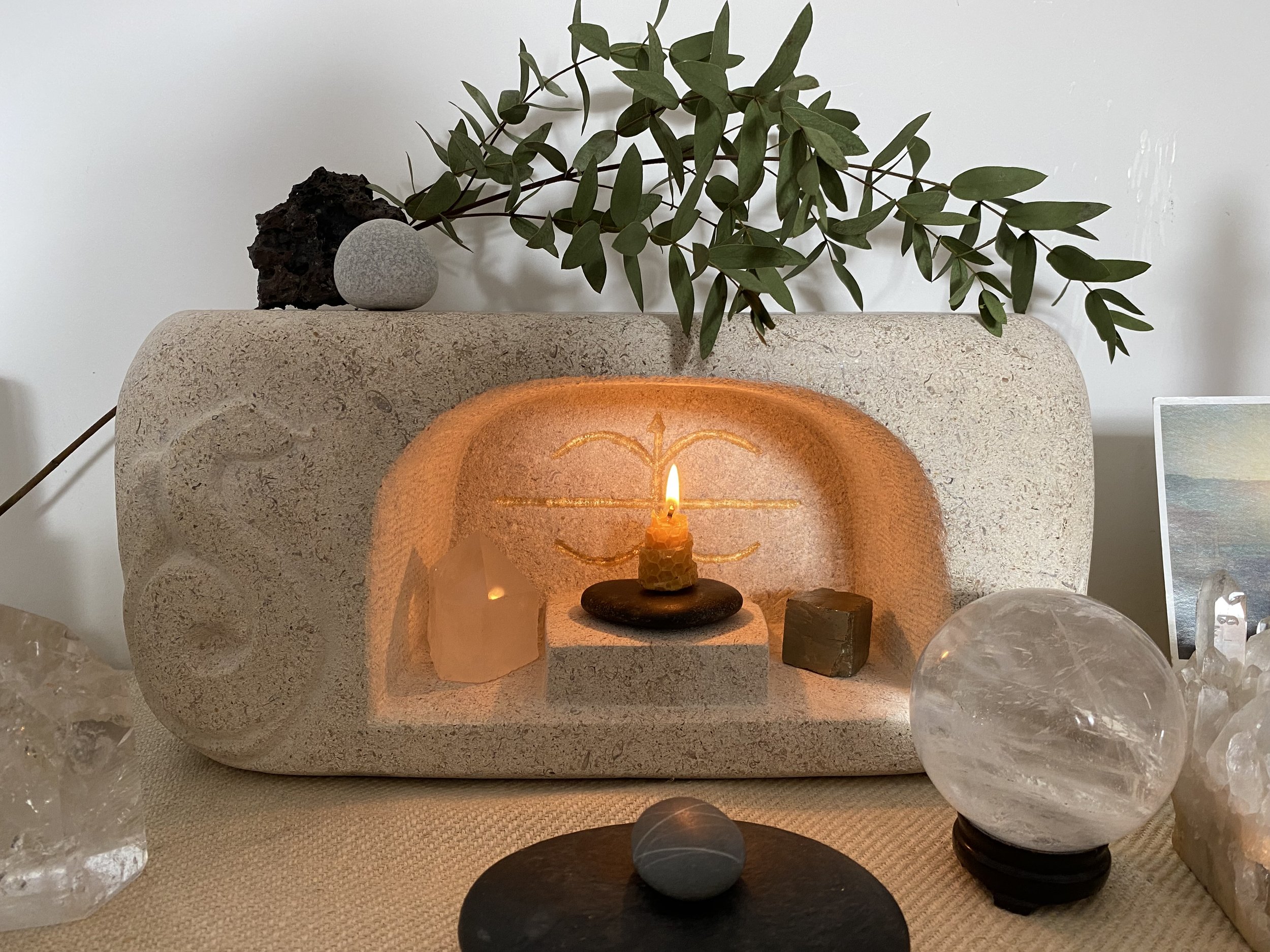 Decorative altar with a lit candle, surrounded by smooth stones, volcanic rock, and crystals, with a green leafy plant on top.