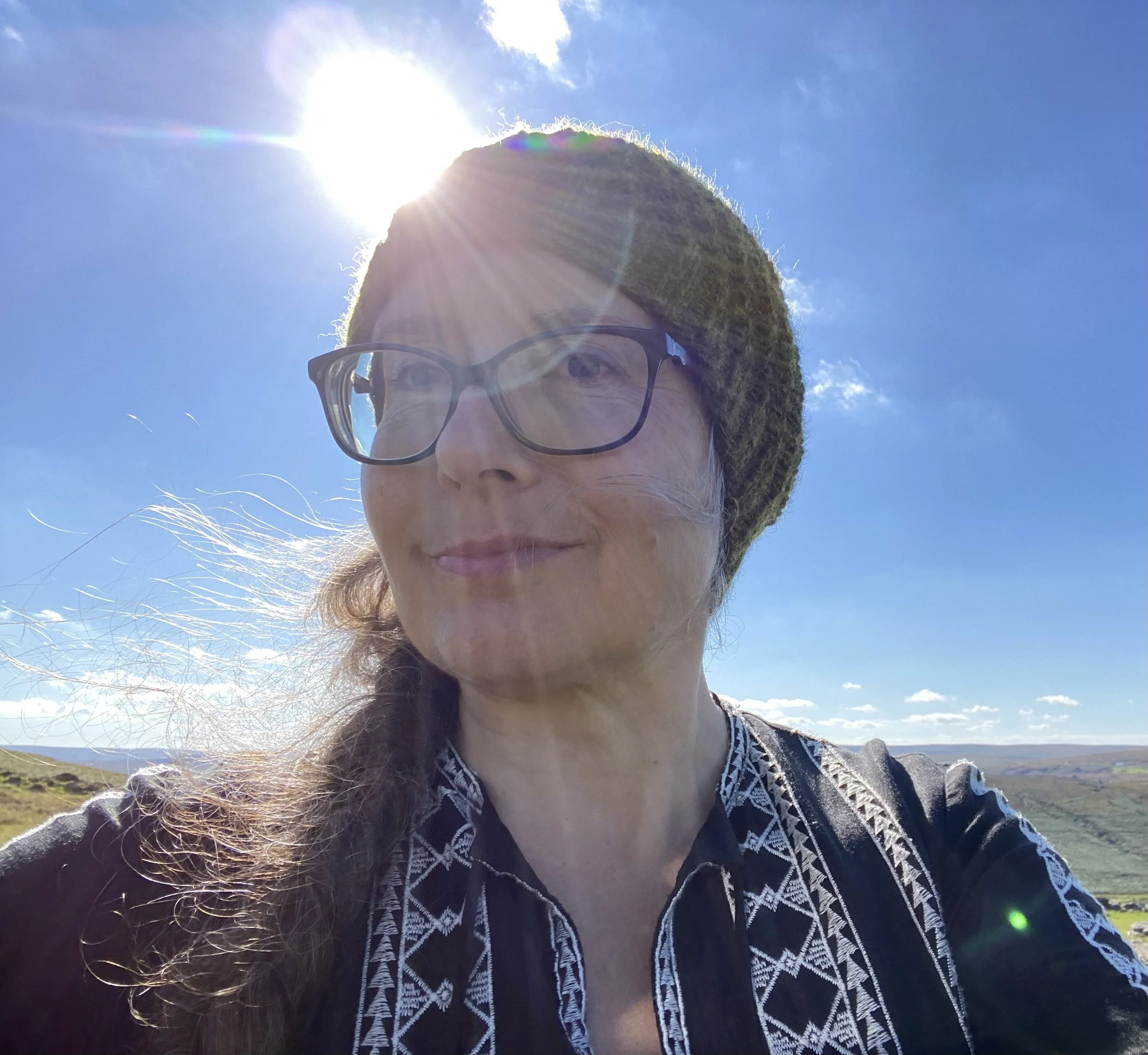 A woman wearing glasses and a knitted hat, standing outside on a sunny day with a blue sky and some clouds in the background.