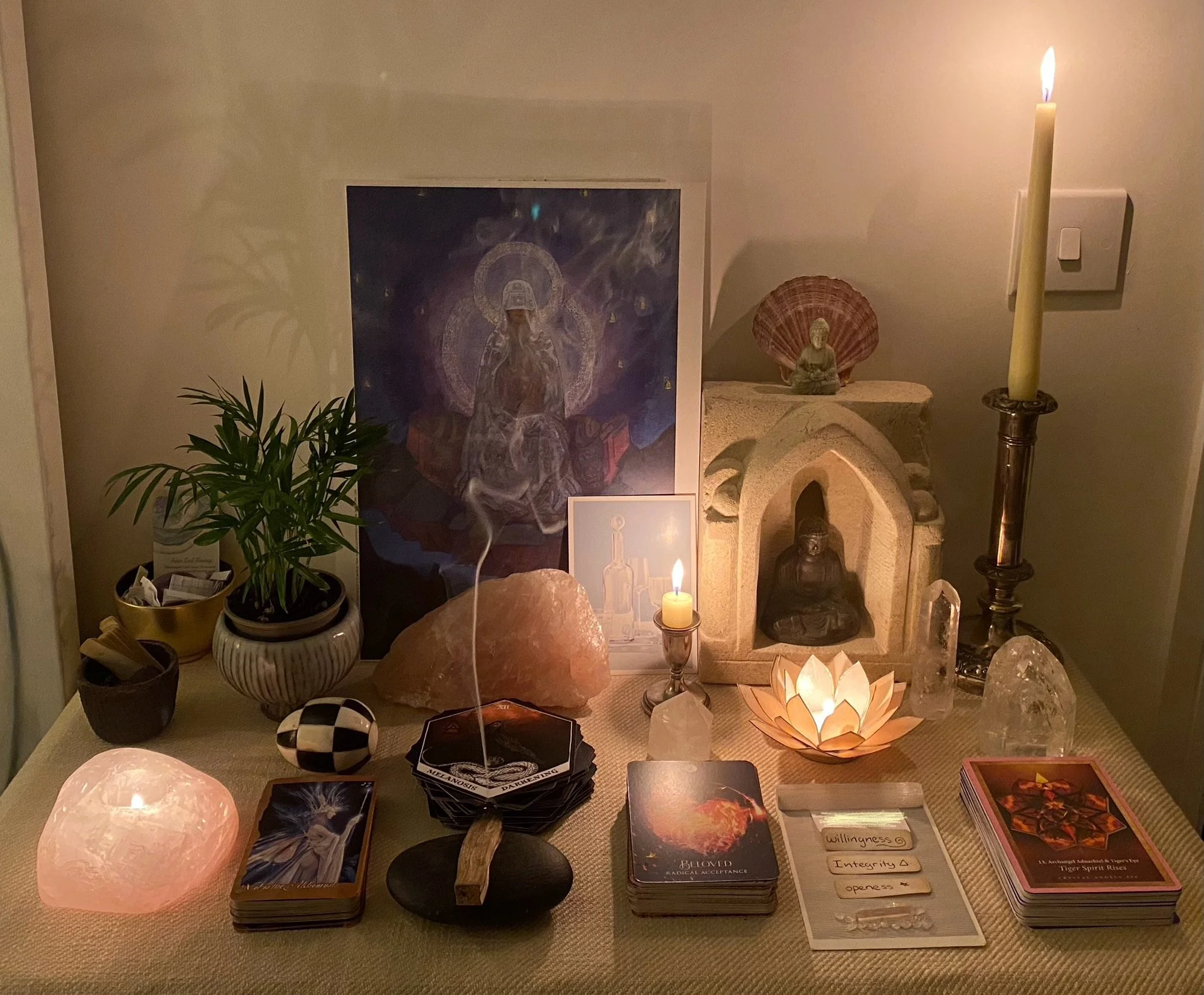 A spiritual altar with a Buddha statue, candle, plants, crystals, tarot cards, and spiritual items, with consciousness-related words written on a paper in front.