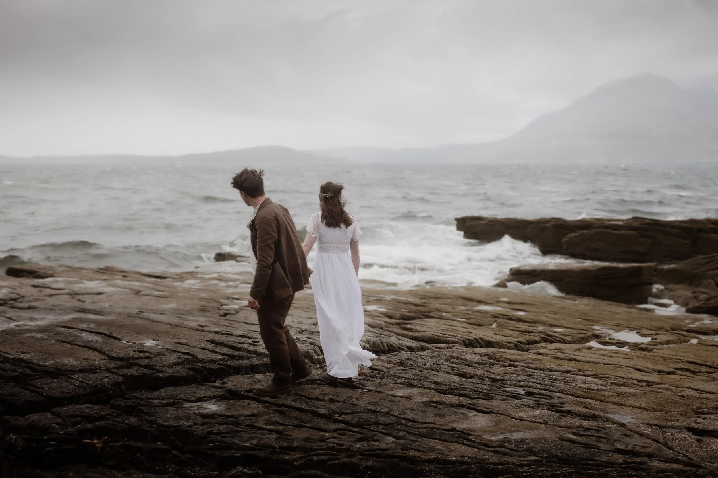 A couple holding hands walking along a rocky shoreline on a cloudy day, with the ocean and mountains in the background.