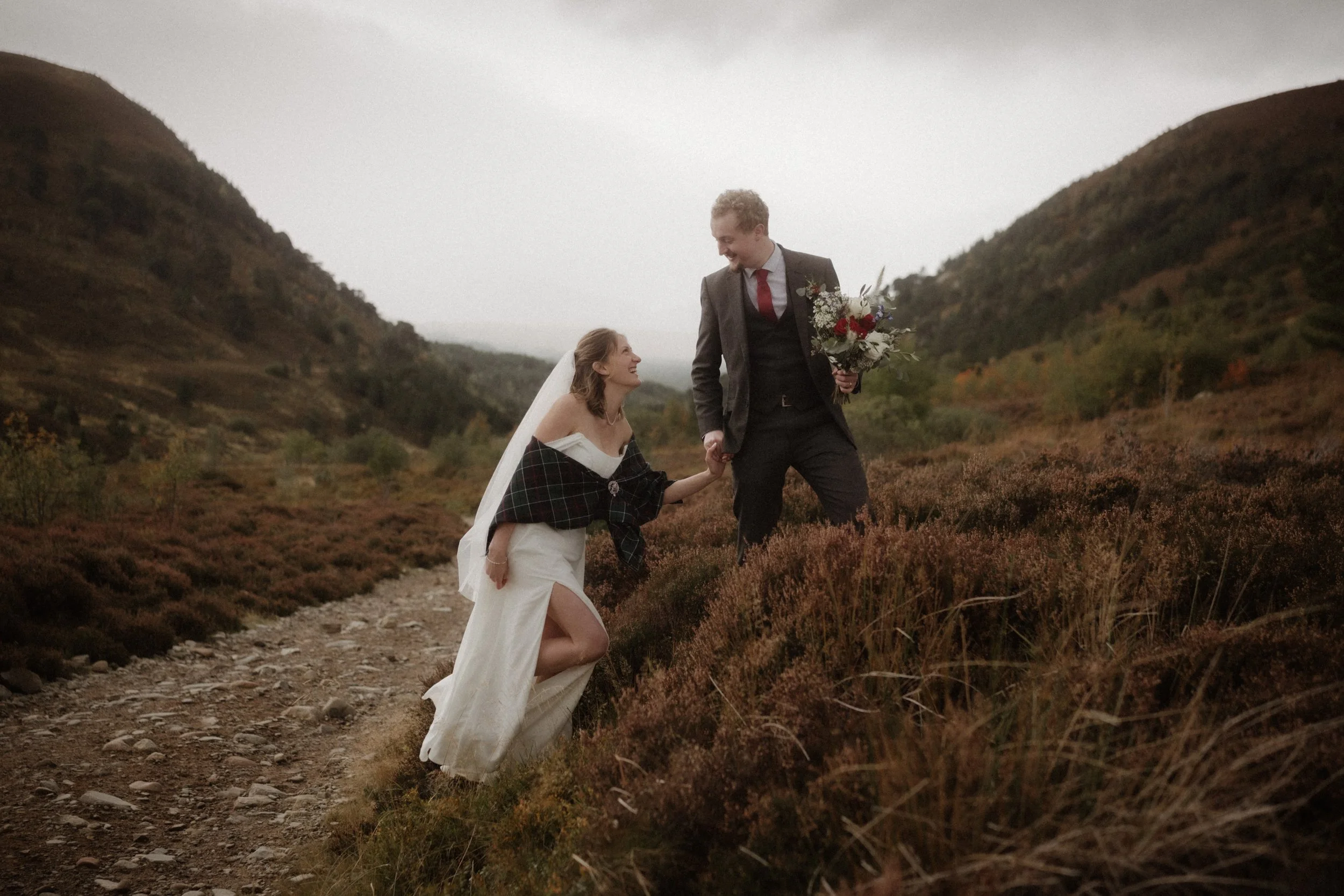 A bride and groom holding hands on a mountain trail with rolling hills in the background, the bride in a white dress and veil, the groom in a dark suit holding a bouquet, both smiling.