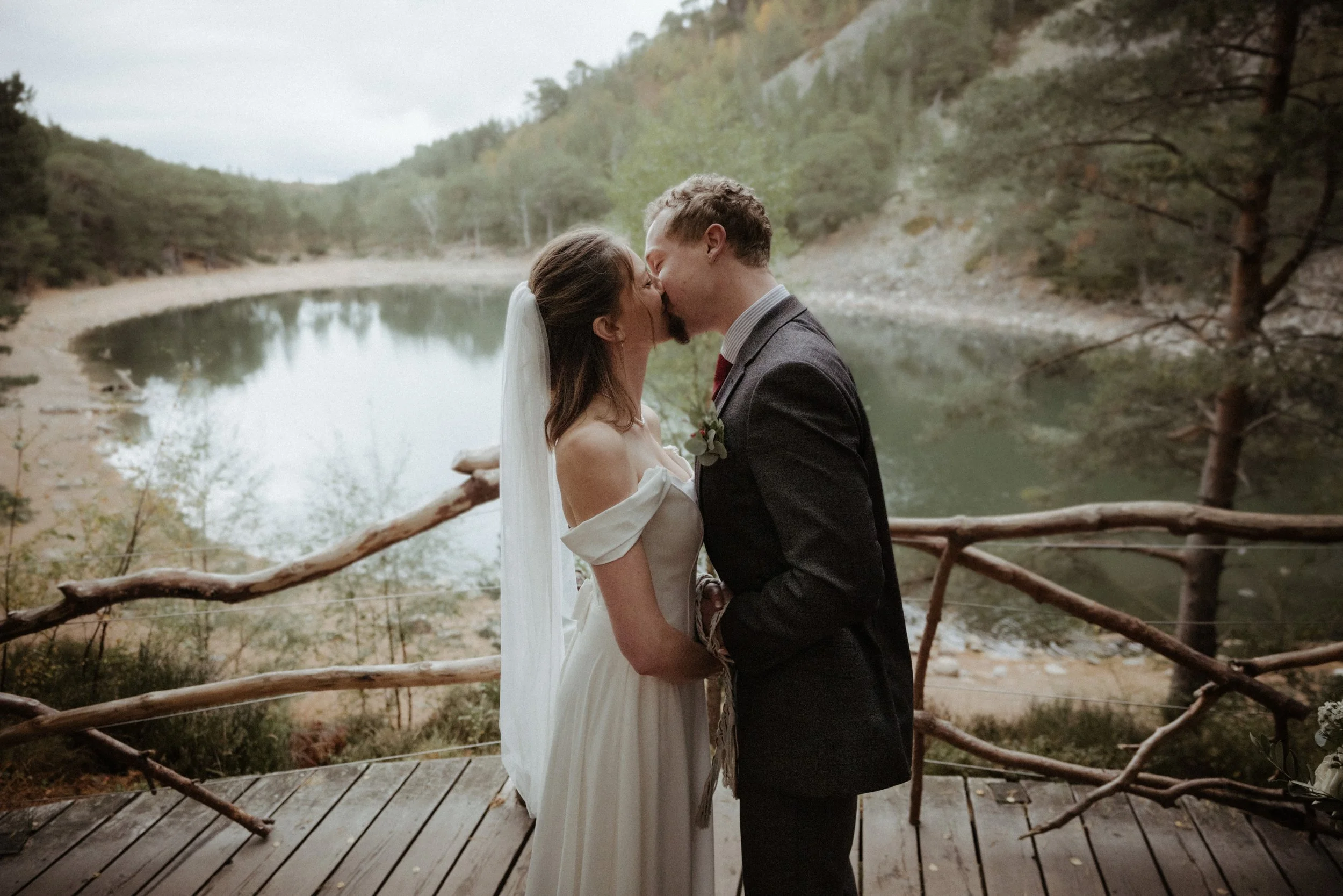 A bride and groom kissing on a wooden deck overlooking a lake surrounded by trees.