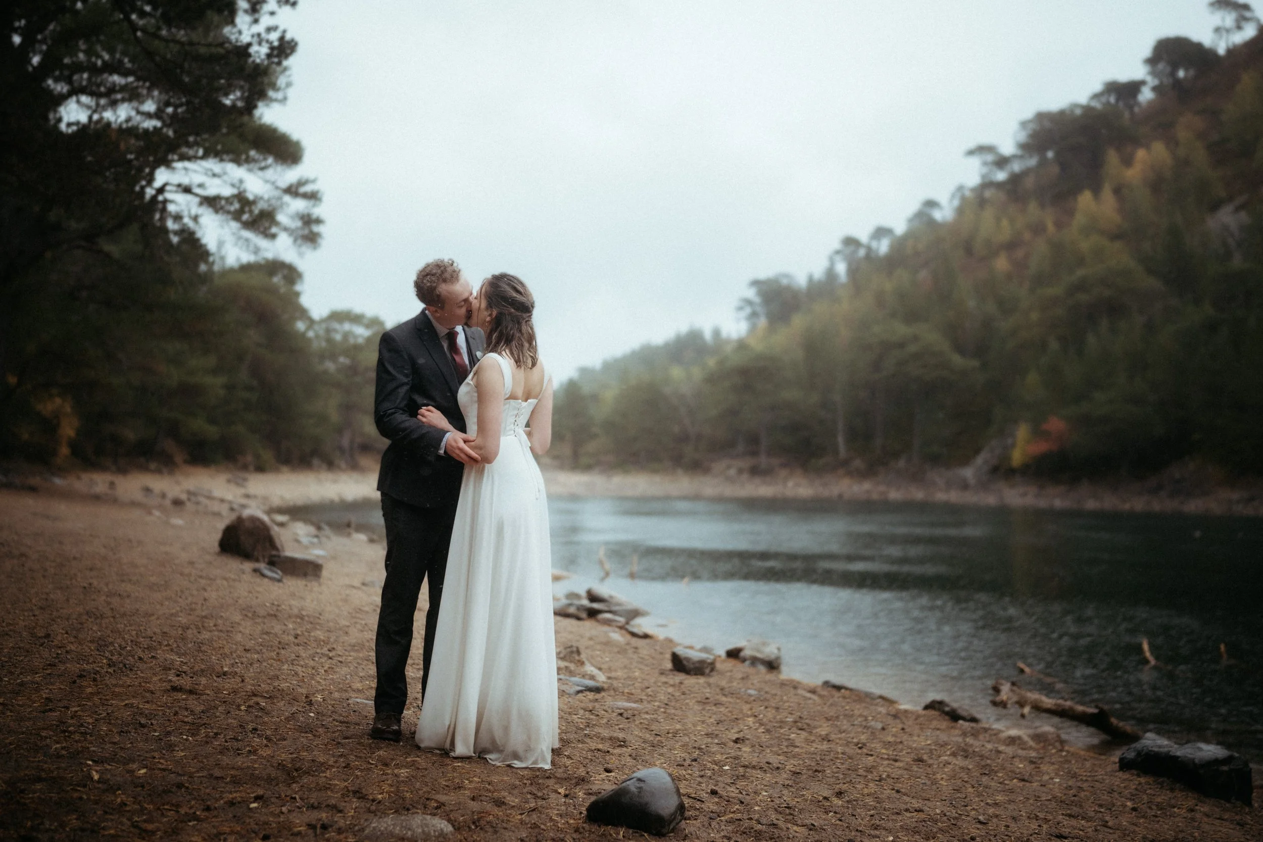 A couple dressed in wedding attire sharing a kiss beside a river surrounded by trees in Aviemore