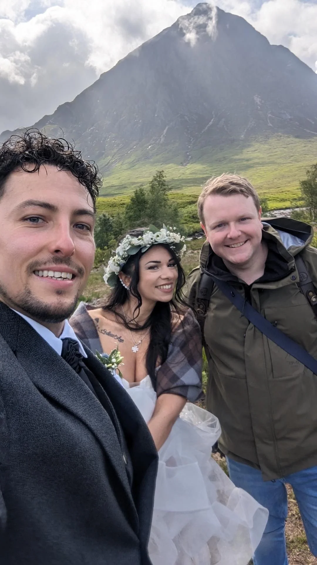 Three friends taking a selfie outdoors in front of a mountain with green slopes and a cloudy sky. One person is dressed in a wedding outfit with a flower crown, another in a jacket, and the third in a suit.
