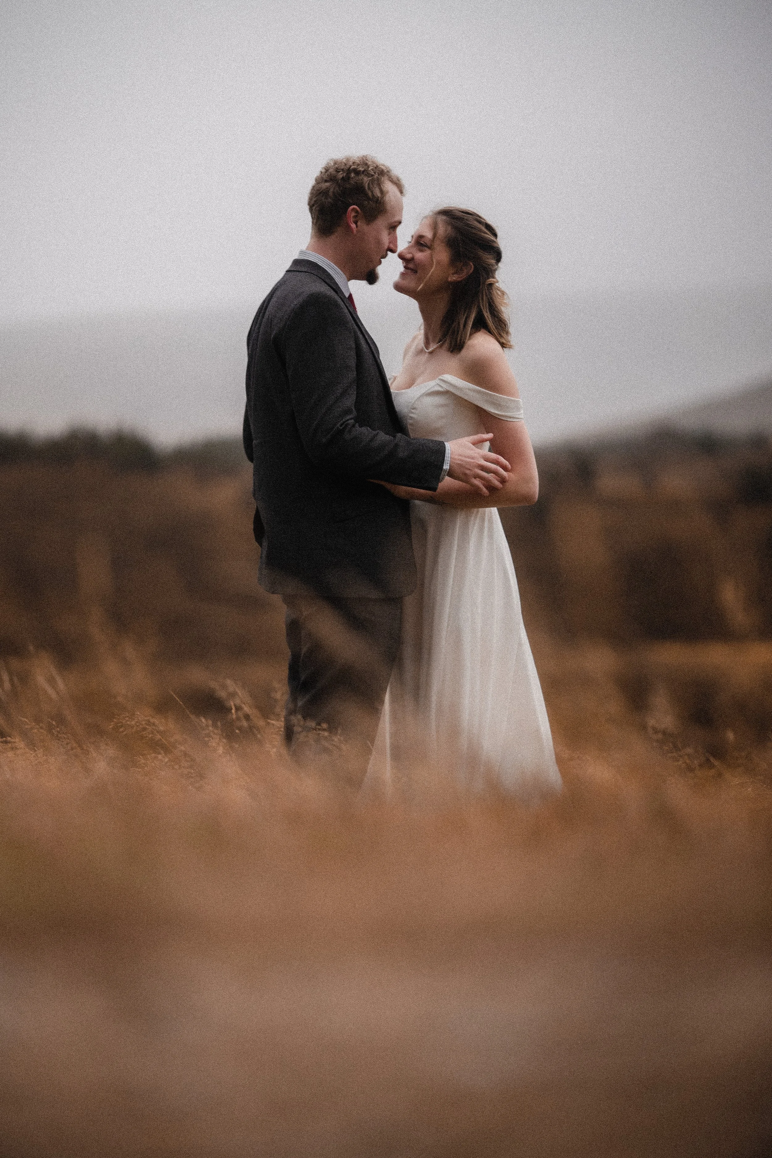 A man in a dark suit and a woman in a white dress stand close together outdoors, embracing and smiling at each other with a blurred natural landscape in the background.