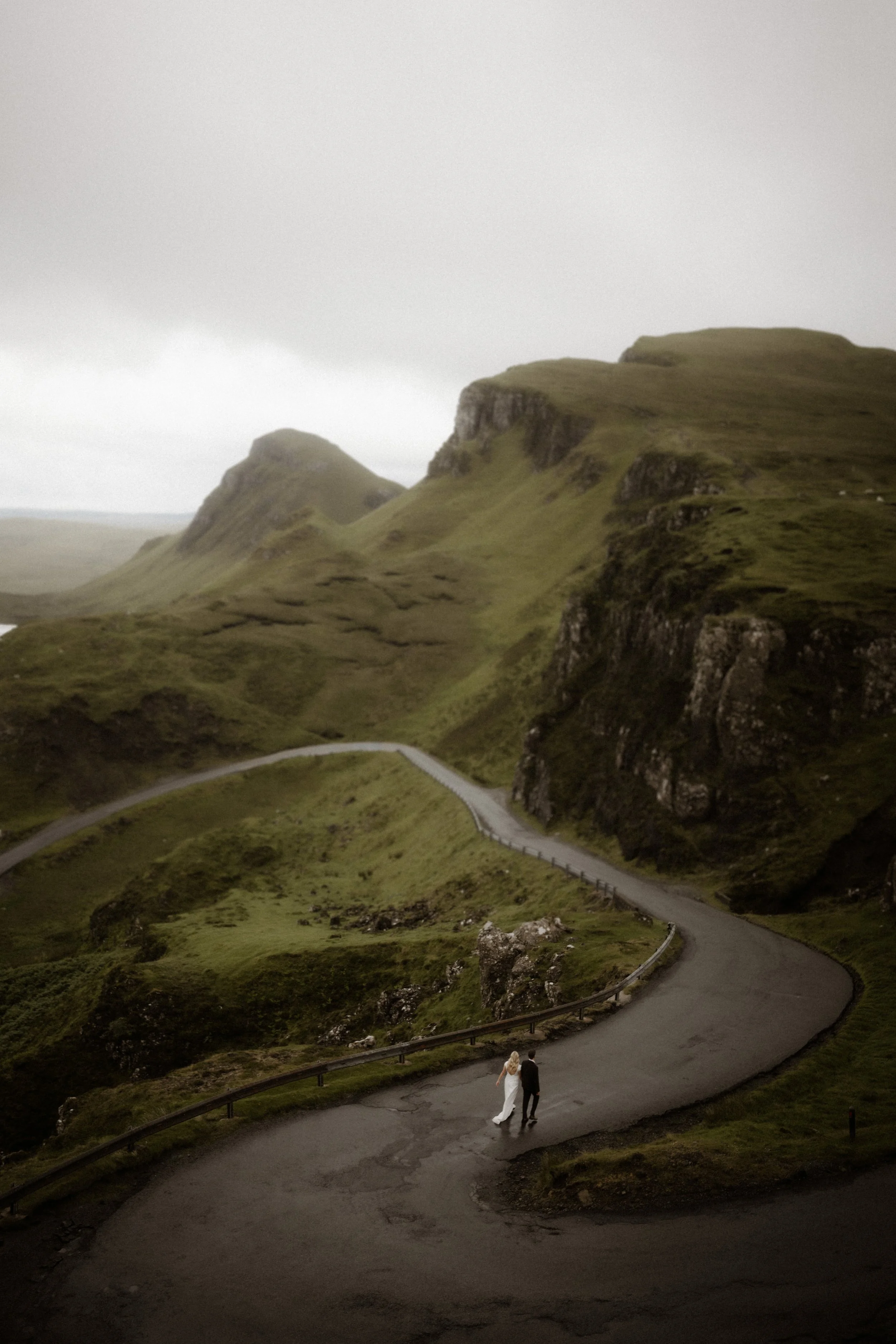 A bride and groom walking along a winding mountain road surrounded by lush green hills and rocky cliffs under a cloudy sky.